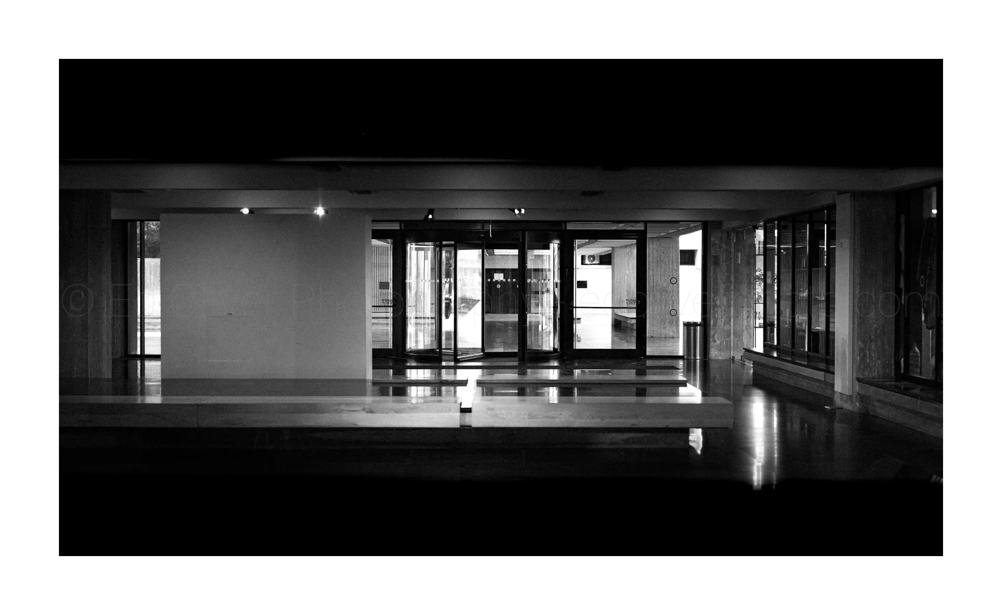 The interior of a modern building lobby with glass doors, wooden accents, and reflections on the polished floor in black and white.