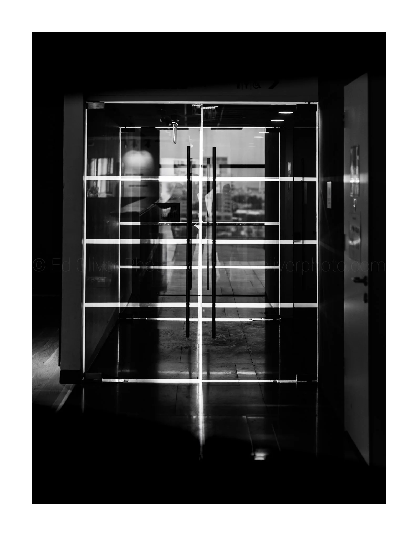 Black and white photo of a modern glass door with interior lighting reflections, leading to a building interior with a cityscape visible outside.