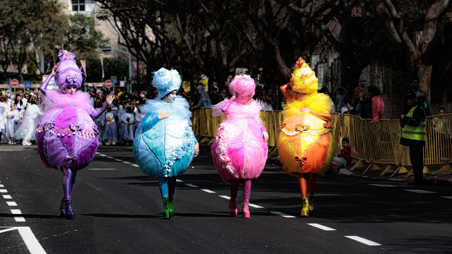 Four people dressed in colorful, elaborate costumes resembling birds or fantasy creatures walking in a parade on a city street, with spectators and security guards watching from behind a barrier.