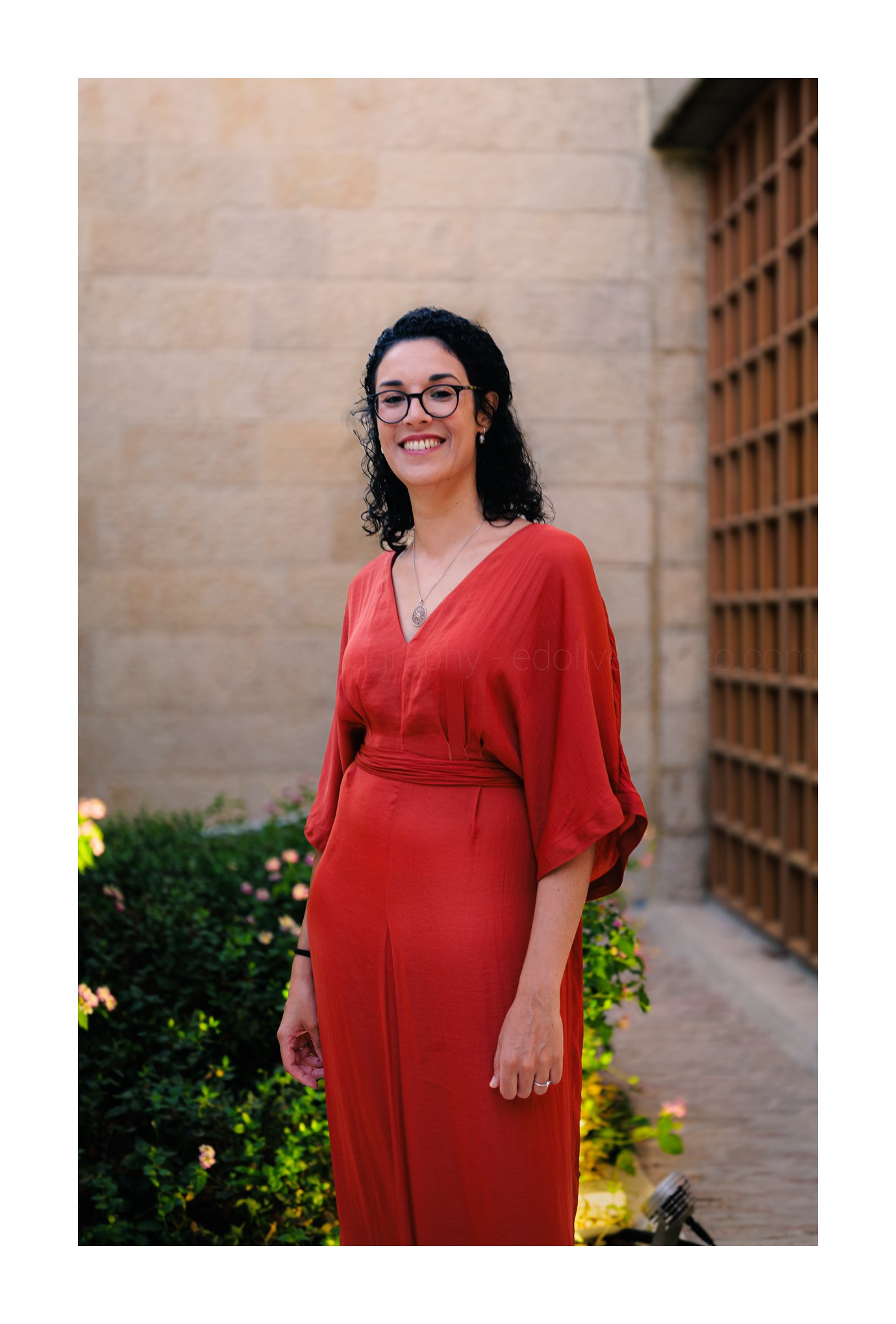 A woman with black curly hair, glasses, and a red dress standing outdoors in front of a stone wall and lattice structure, smiling.