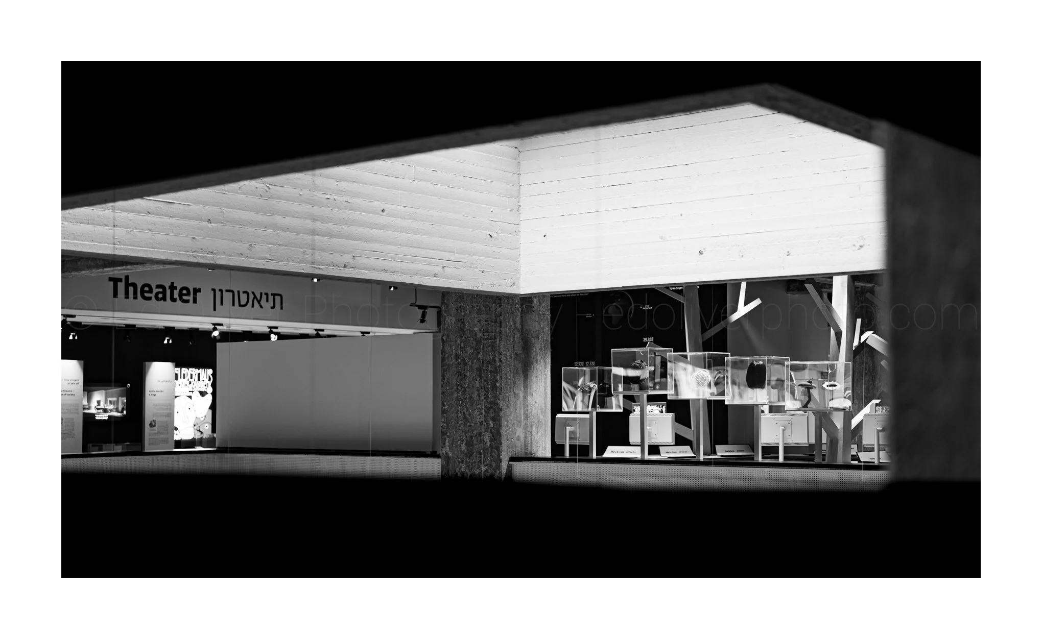 Black and white photo of an indoor exhibit space with a sign that says 'Theater' in English and Hebrew, displaying various objects including electronic devices and art pieces.