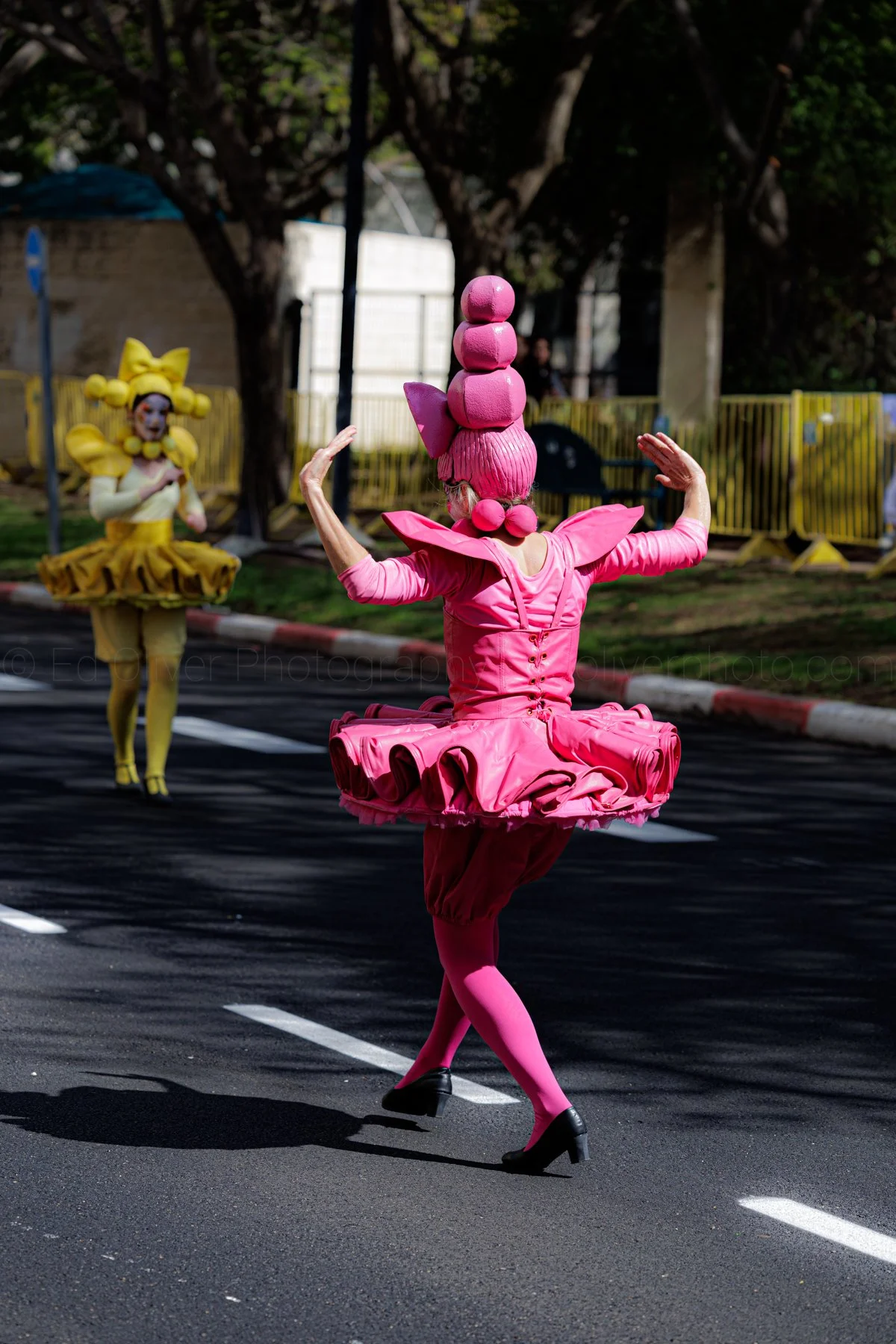 A person dressed in a bright pink, cartoonish costume with a ball-shaped headpiece, flying saucer-like tutu, and high heels, balancing on one foot while dancing on a street. In the background, another person is dressed in a yellow costume with a larg