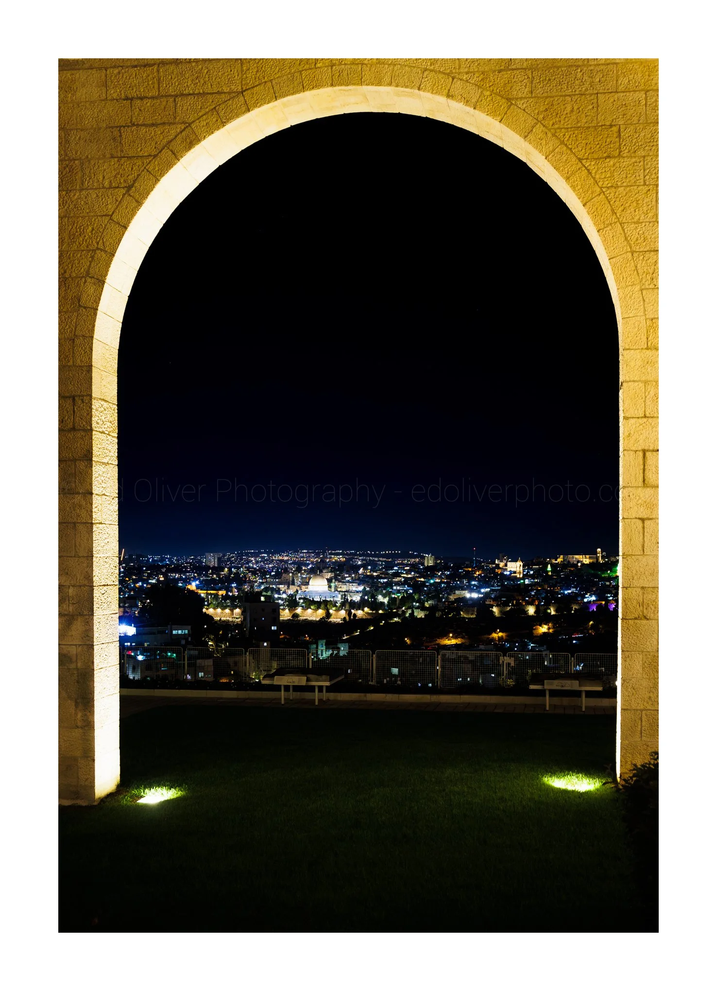 Night view of a cityscape through an arched stone window with illuminated walls and green ground lights.