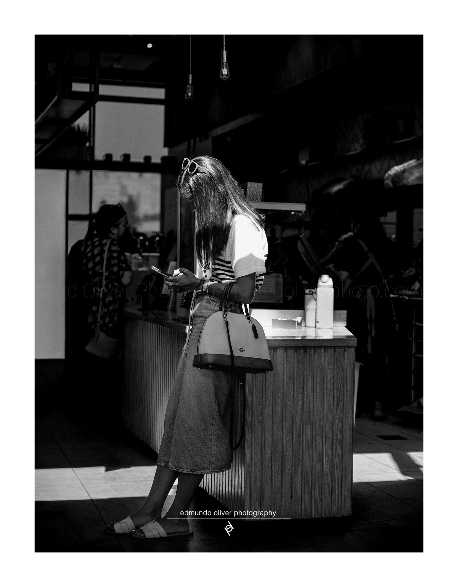 A young woman wearing a striped t-shirt and a long denim skirt is standing at a counter, looking at her phone. She has long hair, sunglasses on her head, and a handbag over her shoulder. Two light bulbs hang from the ceiling, and there are people in 