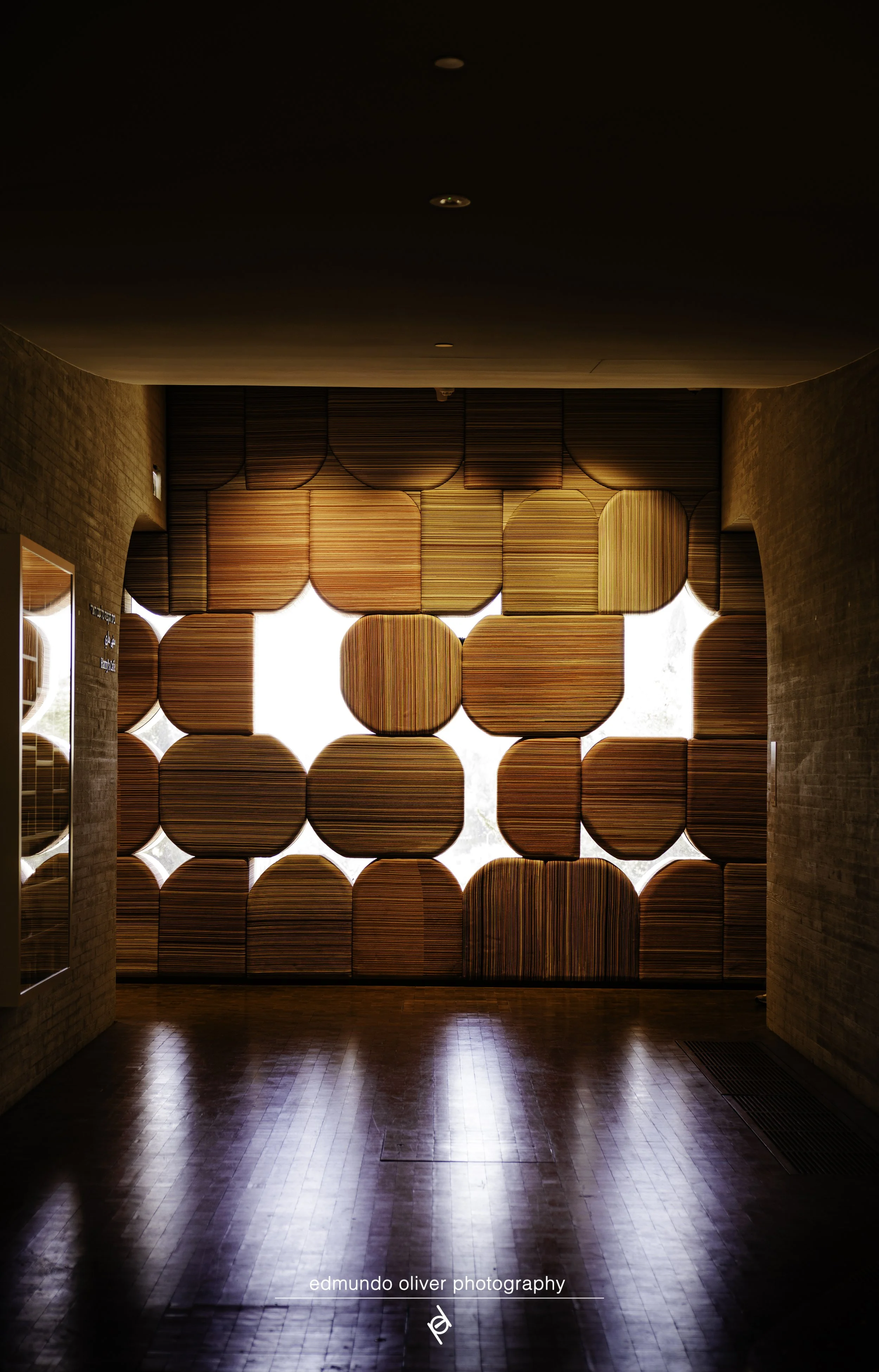 Interior of a building with a polished wooden floor, brick walls, and a decorative wooden screen with circular and semi-circular patterns allowing light to pass through.