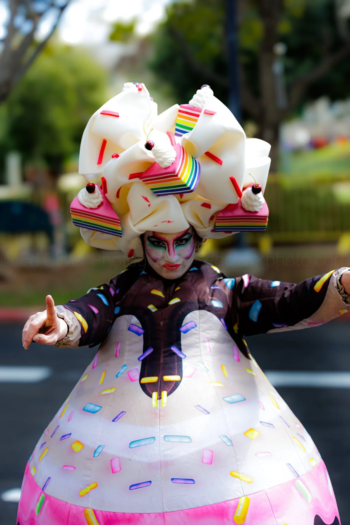 A performer in a colorful, candy-inspired costume with a large, elaborate headpiece resembling frosting and sprinkles, during a parade or festival.