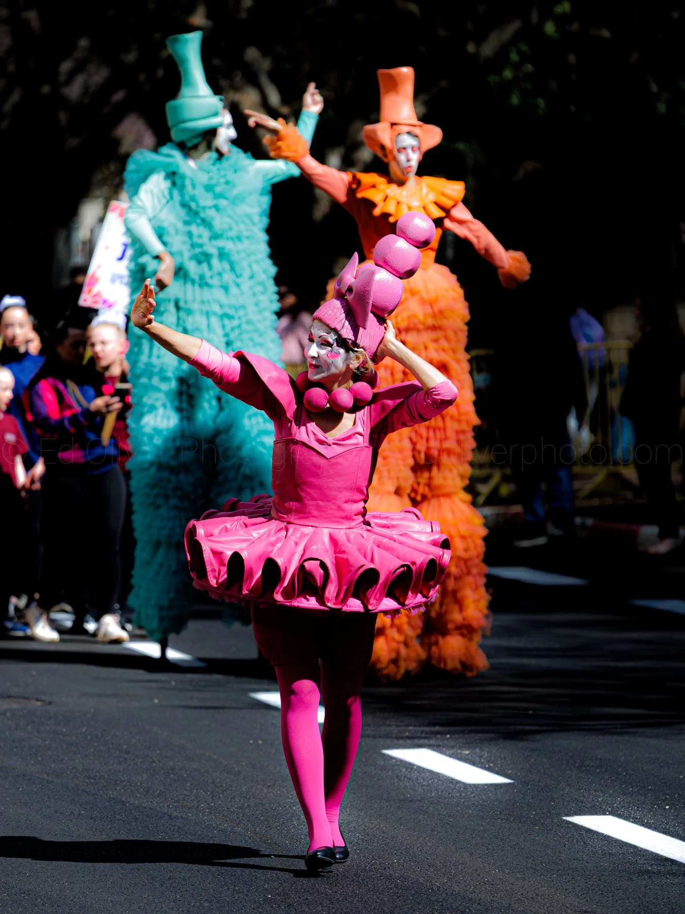 Performer dressed as a pink candy-like character with a tutu and tall pink confection hat, leading a parade on a street.