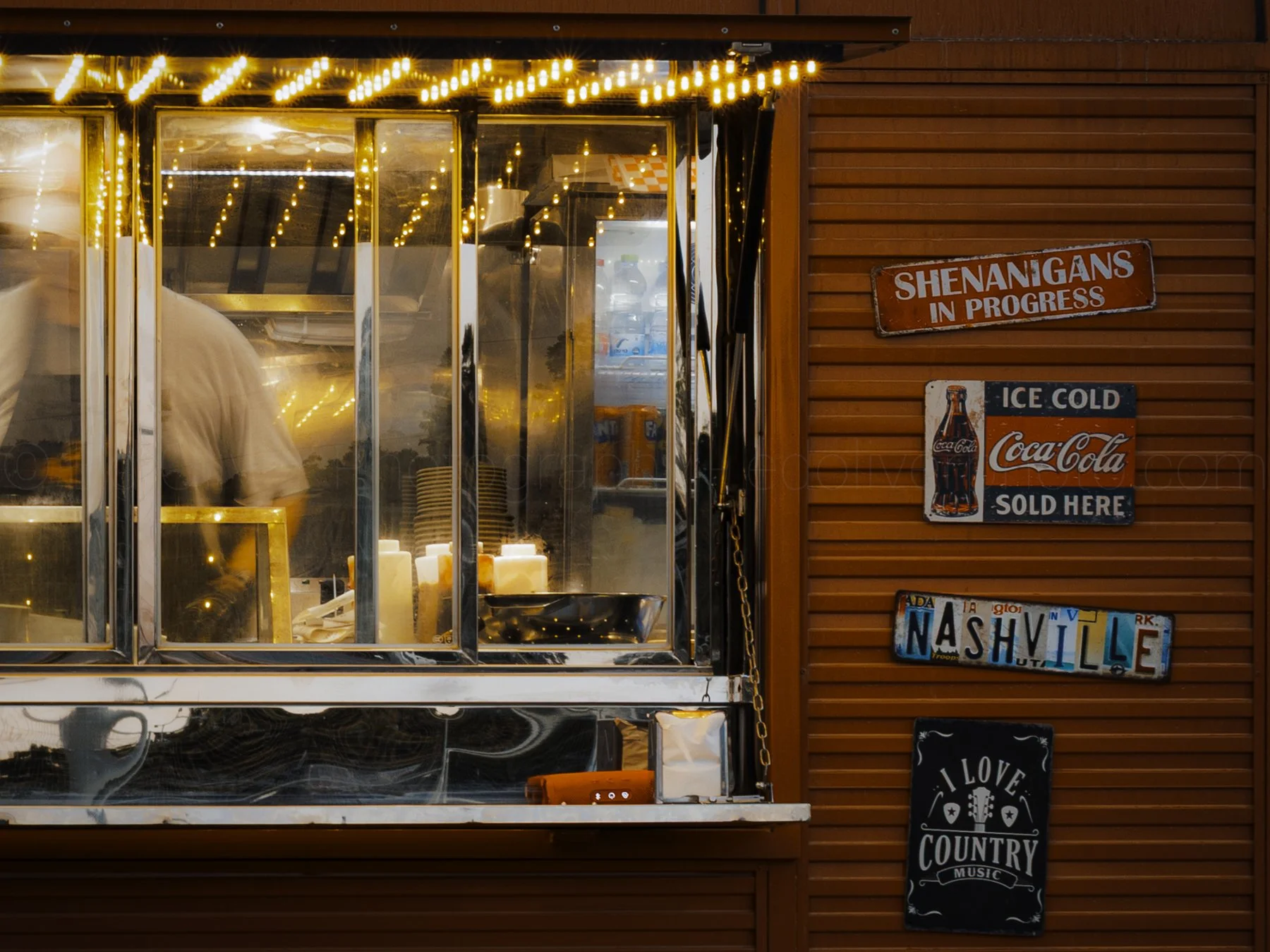 A roadside food stand with warm string lights above a glass window counter, showing a person working inside. The stand has signs including one that says "SHENANIGANS IN PROGRESS" and a Coca-Cola sign indicating ice cold drinks sold here. A Nashville 