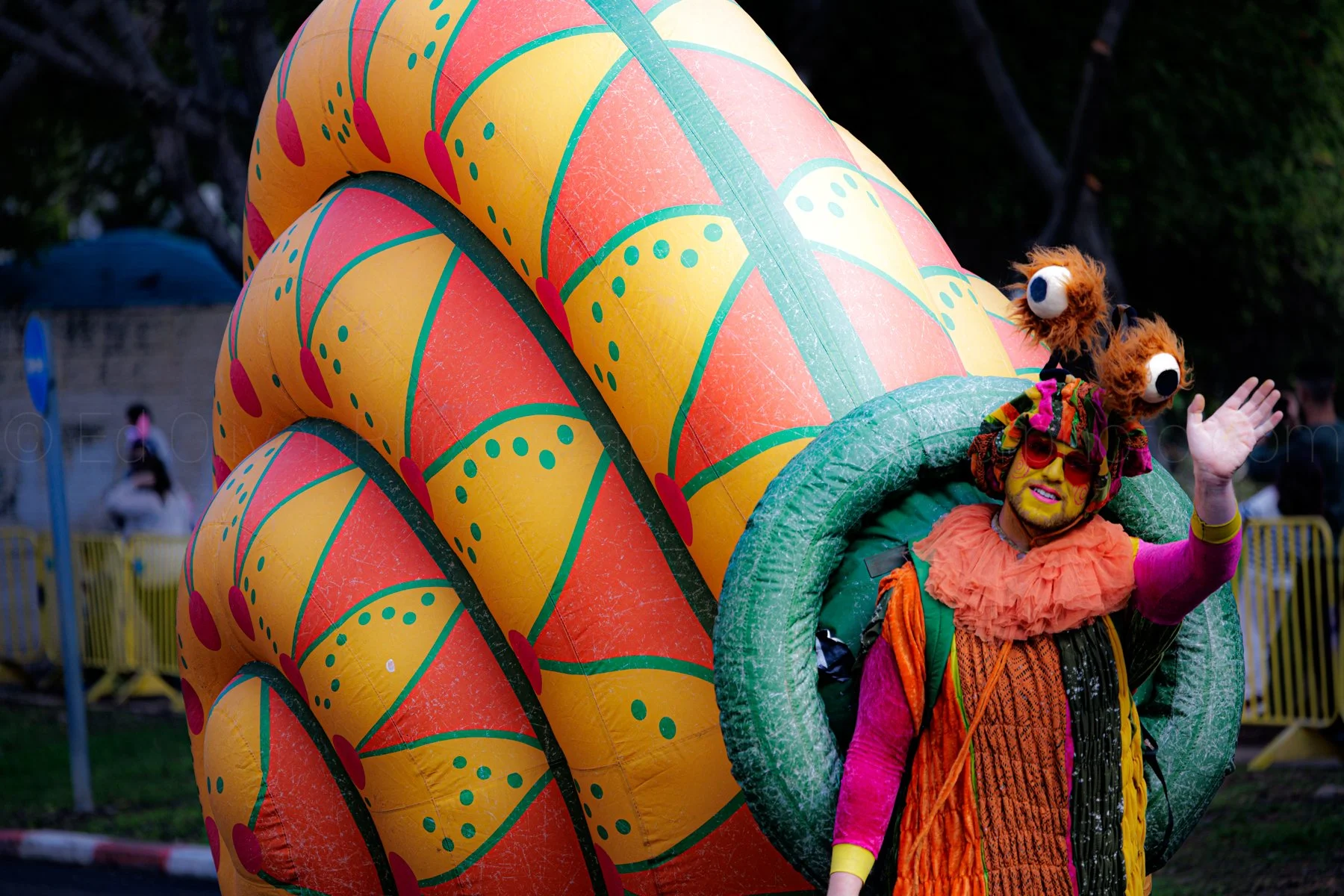 Person dressed in colorful clown costume with orange, pink, and yellow clothing, wearing red sunglasses, waving, standing inside a large, bright decorated prop resembling a pineapple, with an outdoor background and some people and barriers visible.