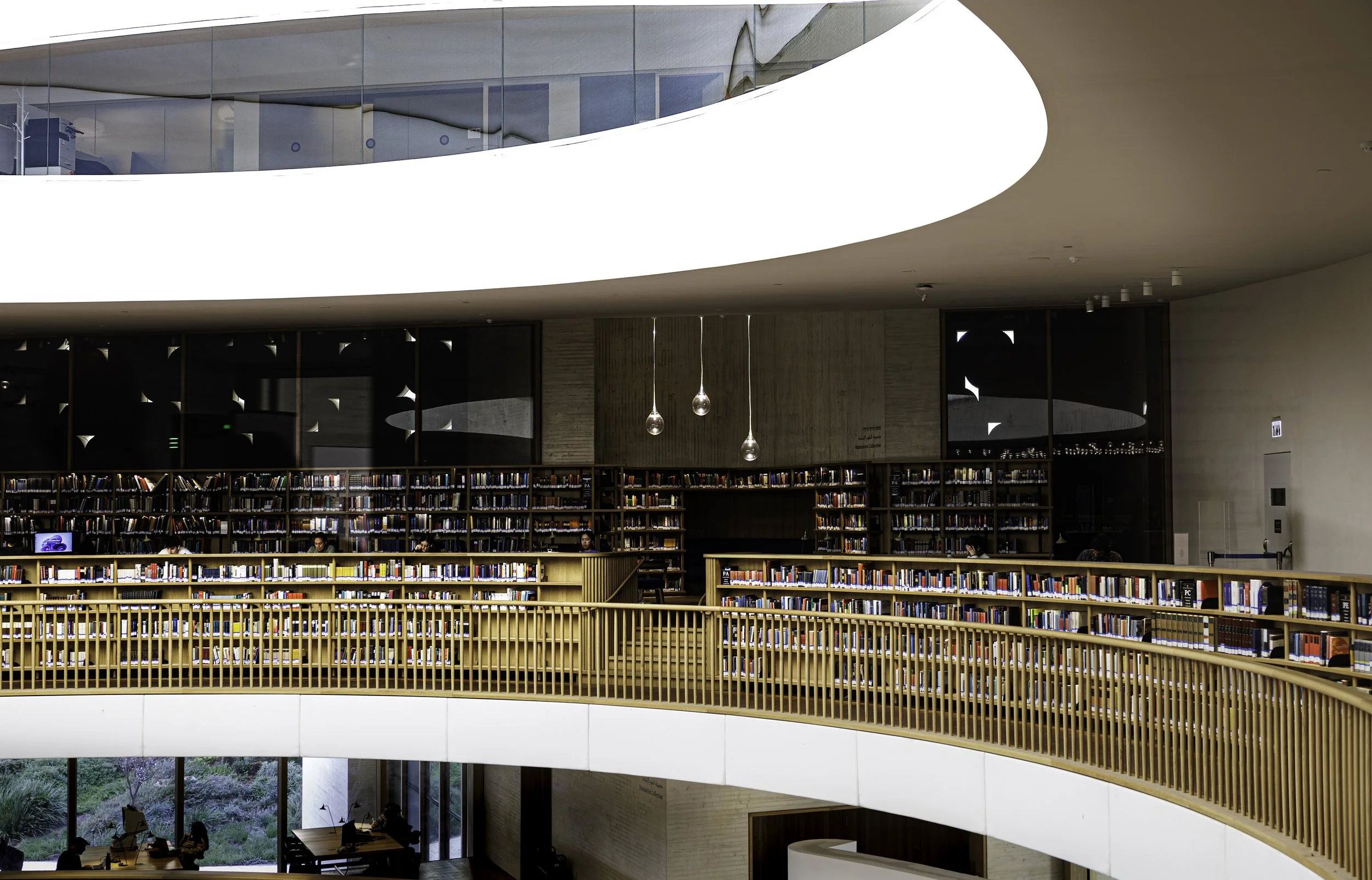 Interior of a modern library with multiple rows of bookshelves, a glass wall at the upper level, and pendant lights hanging from the ceiling. There are people working at desks near a large window with a view of greenery outside.
