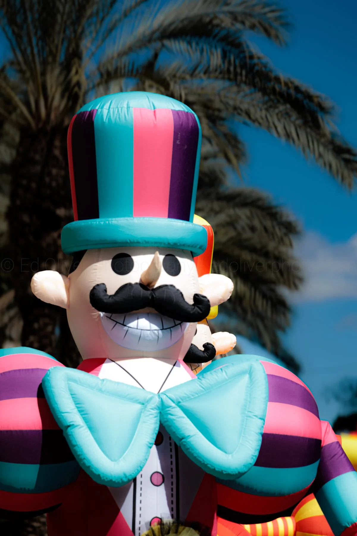 Colorful inflatable clown with a large teal and pink striped top hat, black mustache, big smile, and wearing a teal bow tie, against a backdrop of palm trees and blue sky.