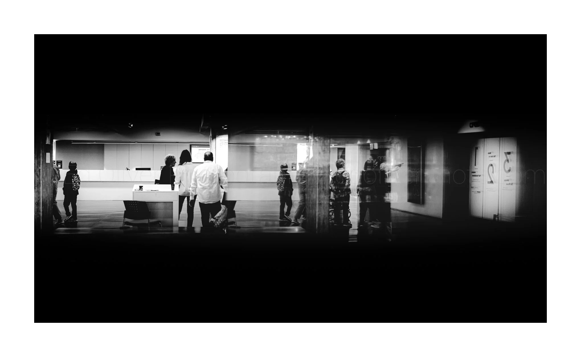 People waiting at a check-in counter area in an airport terminal, seen through a glass window in black and white.