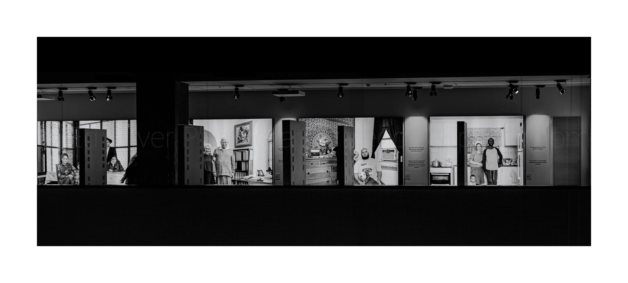 Black-and-white photo of a display showcasing various rooms with people, including children, adults, and elderly individuals, some standing and some seated, inside a museum or exhibit setting.