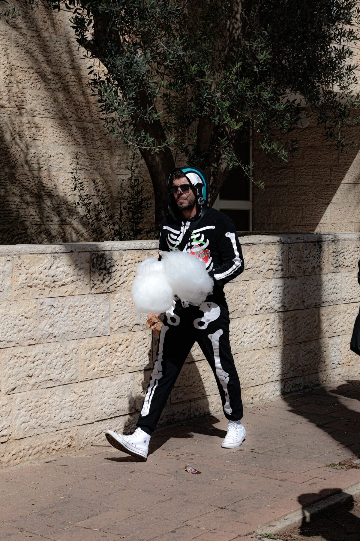 A person dressed in a skeleton costume with bones printed on a black background, holding cotton candy, walking on a sidewalk next to a stone wall and tree.