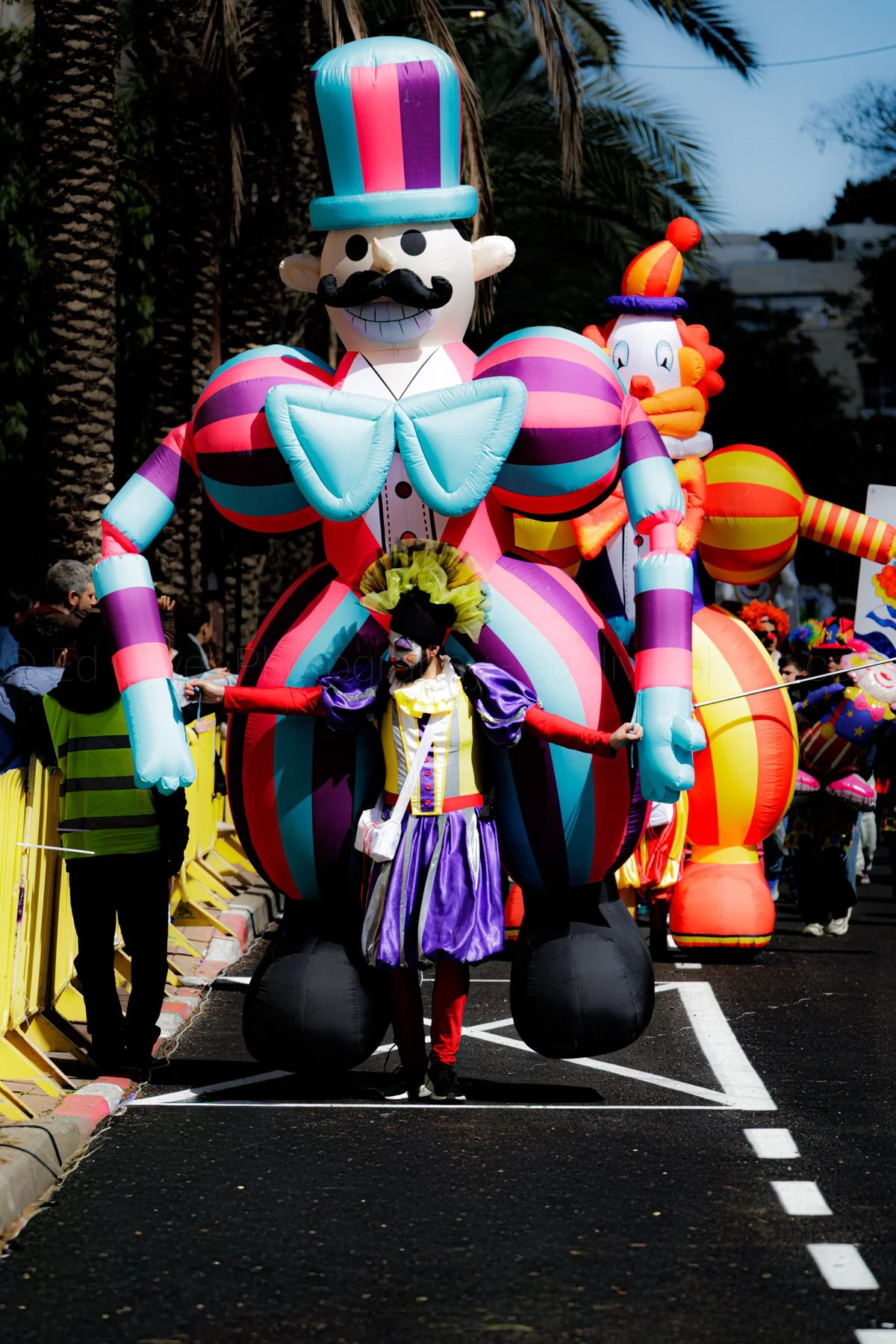 Colorful parade float featuring a large inflatable clown with a top hat, bow tie, and striped outfit, with a person dressed as a clown walking in front.