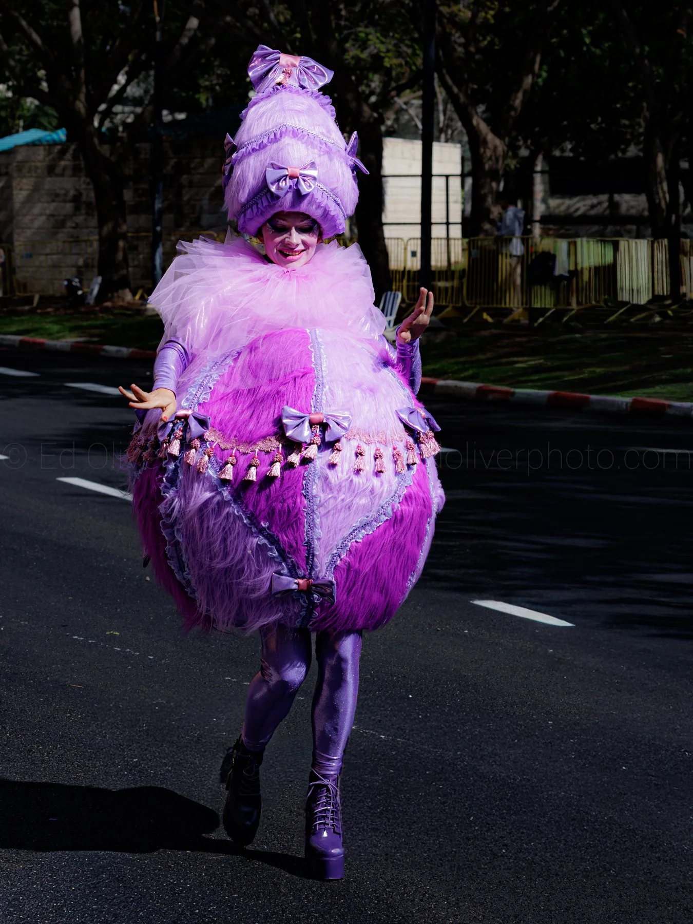 Person in a colorful, elaborate pink and purple costume, resembling a giant, ornate dress with bows and lace, running on a street.