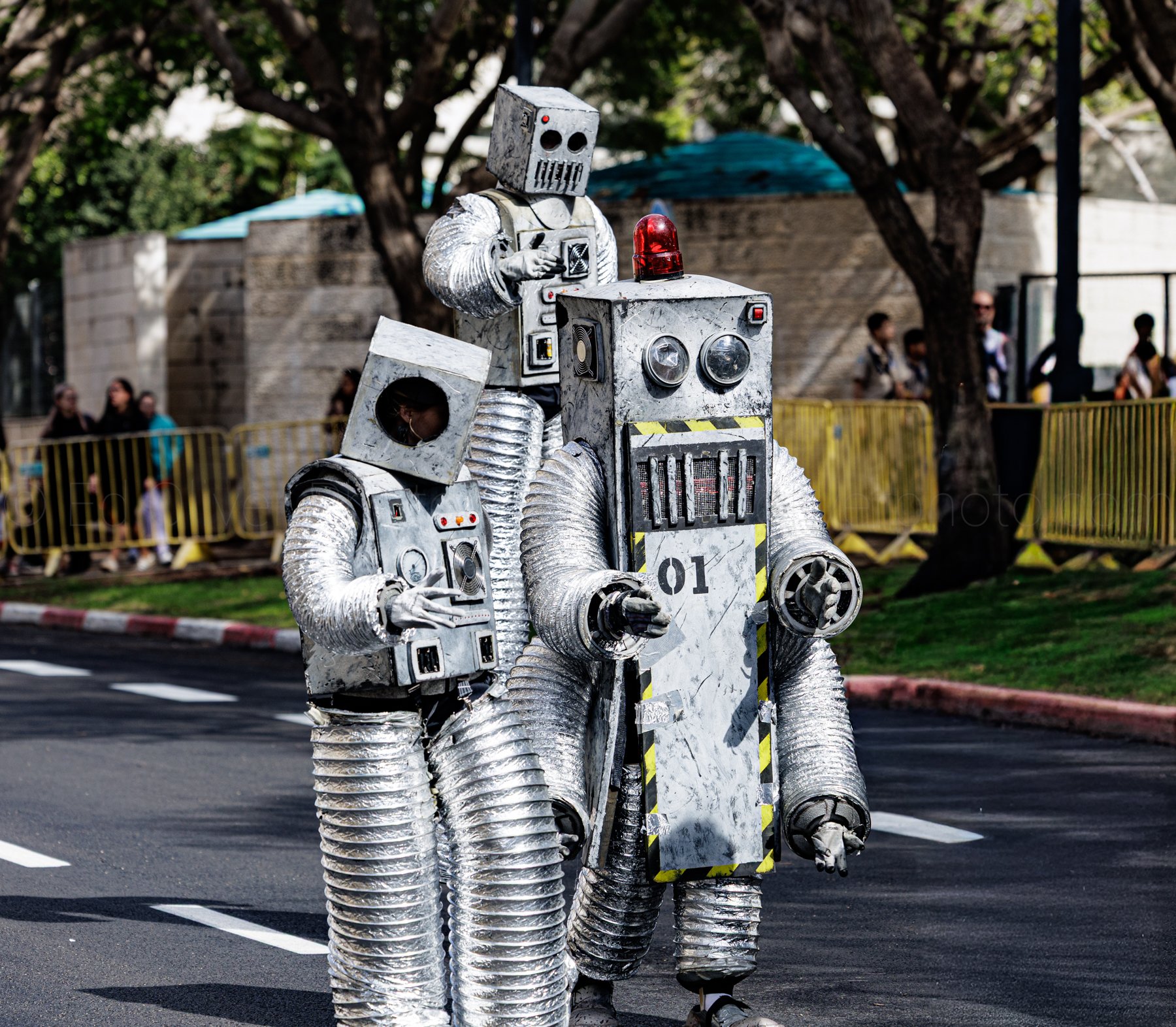 Three people dressed in silver robot costumes walking on a street during a parade or event, with spectators and trees in the background.