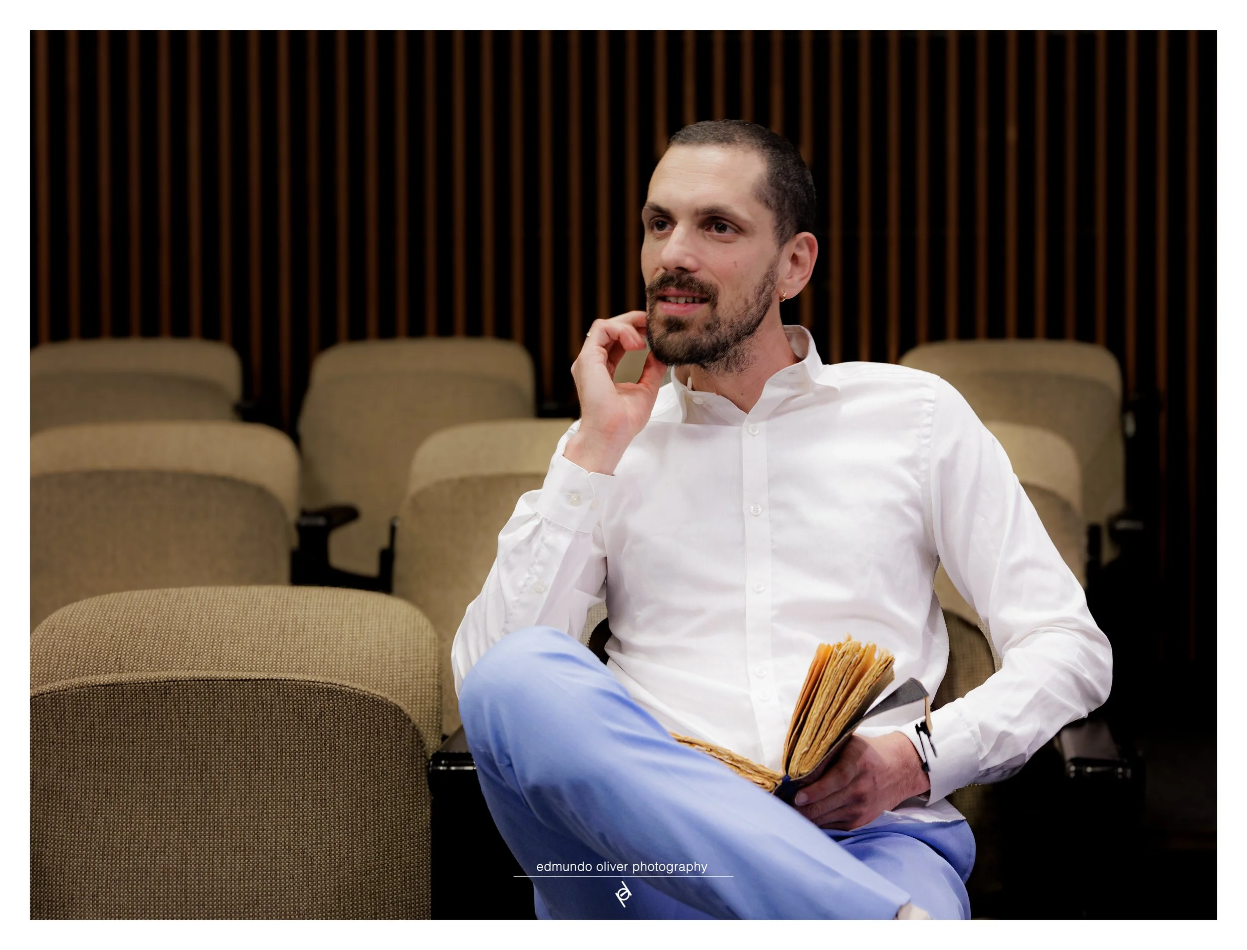 Portrait of a man with short dark hair and a beard wearing a white shirt seated in a room with beige chairs and dark wooden vertical paneling, holding an open book and touching his face with his right hand.