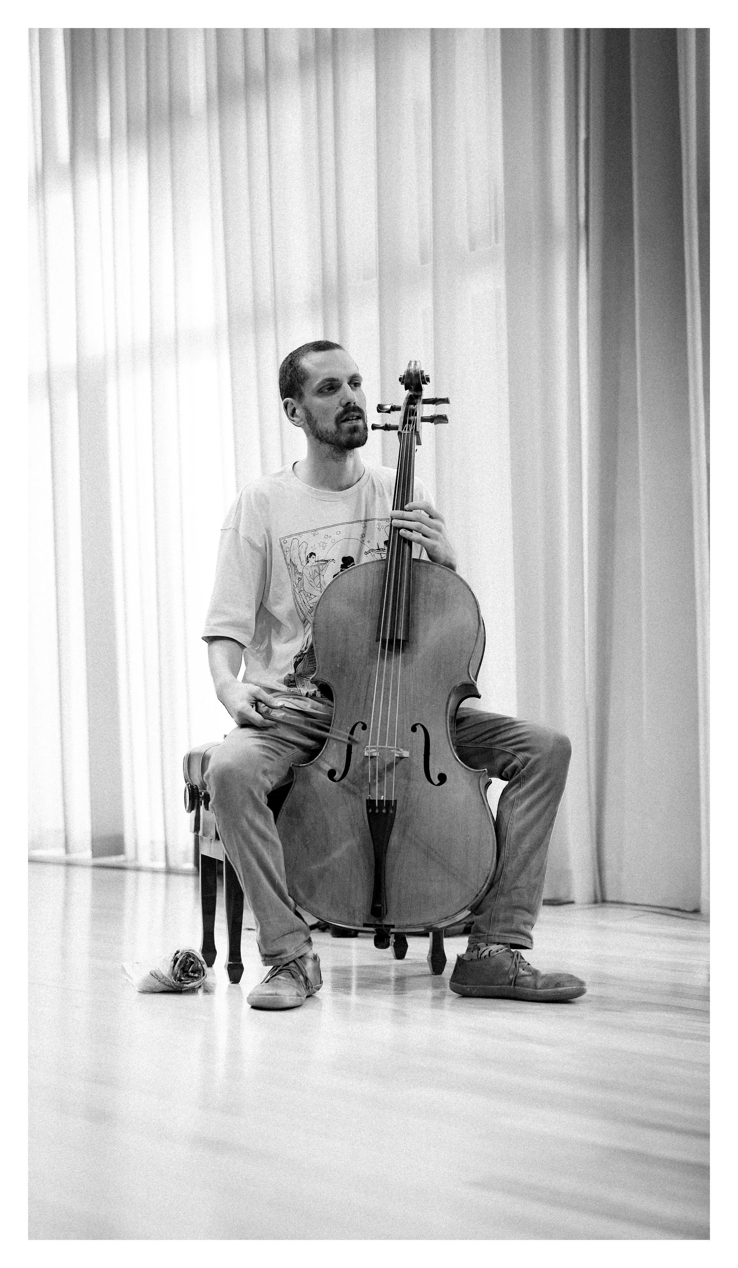 Portrait of a man sitting on a chair and playing a cello in a room with tall curtains in the background.