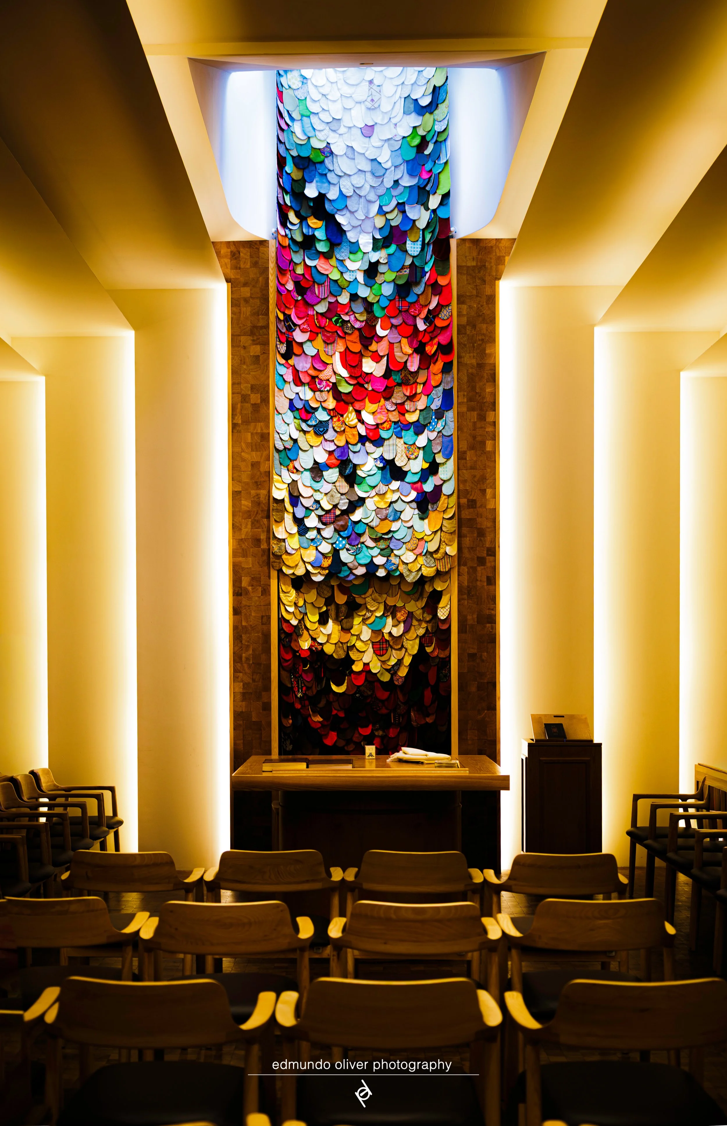 Interior of a room with rows of wooden chairs, a large colorful art piece hanging on the back wall, and a wooden table at the front. The room is lit with warm yellow lighting.