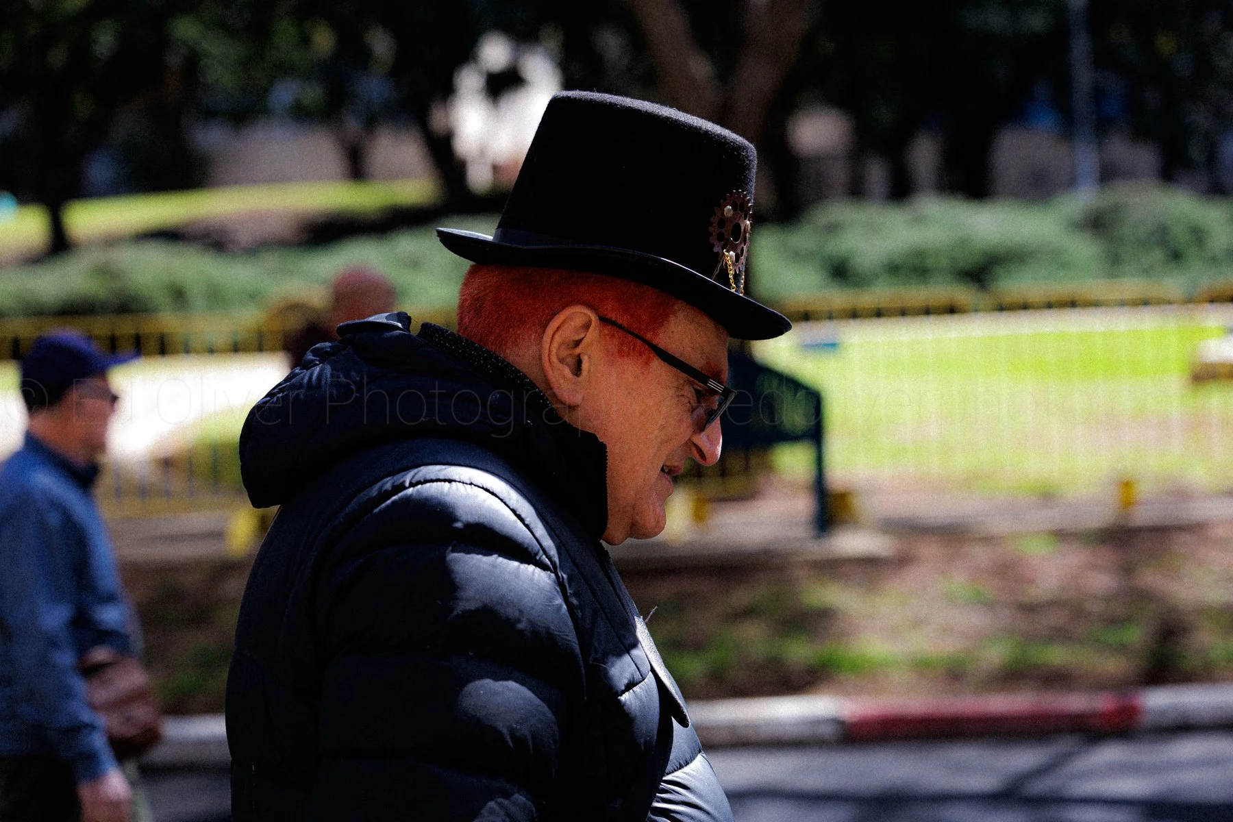A man wearing a black top hat and glasses, smiling, outdoors on a sunny day, with a green park background.