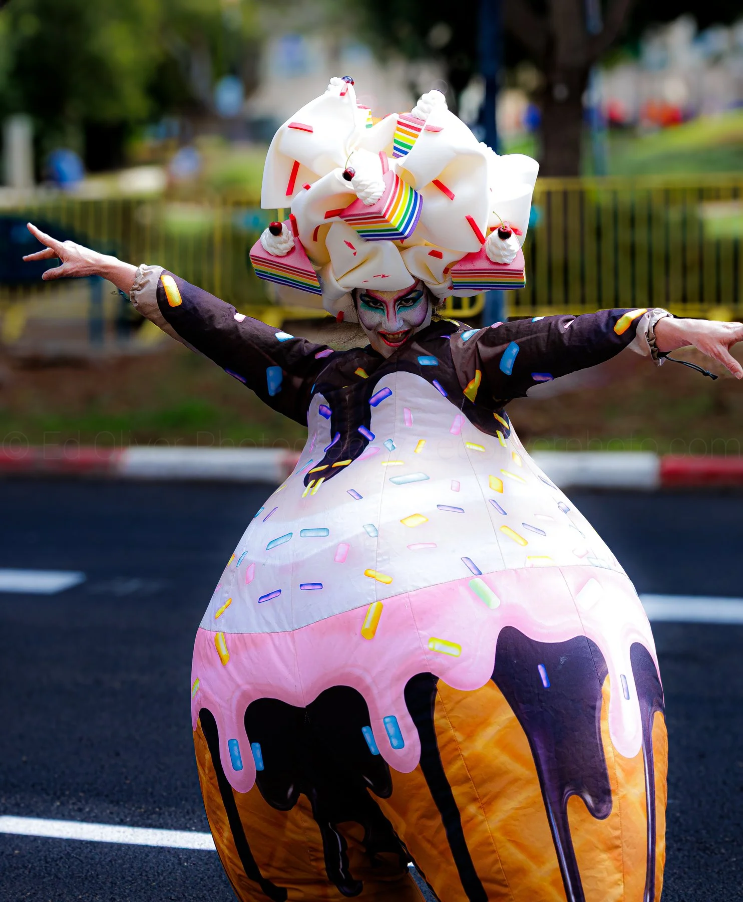 A person dressed in a colorful ice cream-themed costume, with a large headpiece resembling a scoop of ice cream with sprinkles and cherries, poses with arms outstretched in a parade or festival setting.