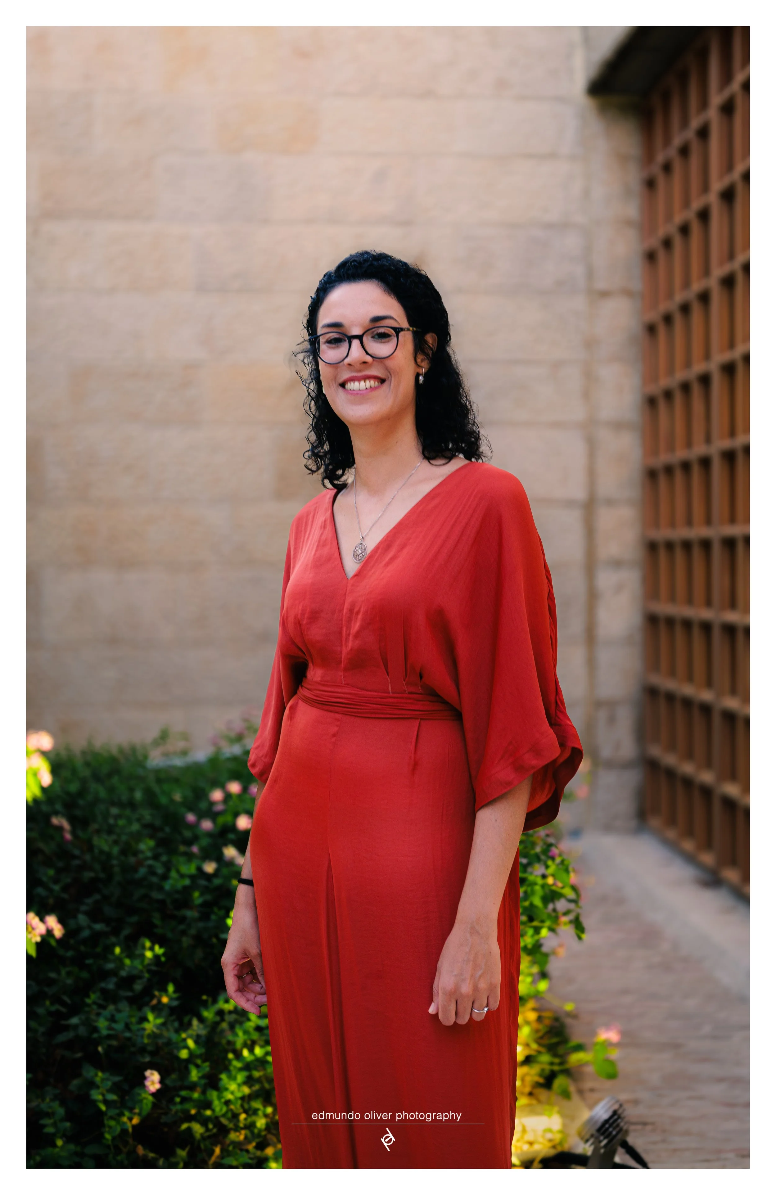 Portrait of a woman with black curly hair, wearing glasses and a red dress, standing outdoors in front of a stone wall and a wooden lattice structure, with flowers and greenery at her feet, smiling at the camera.