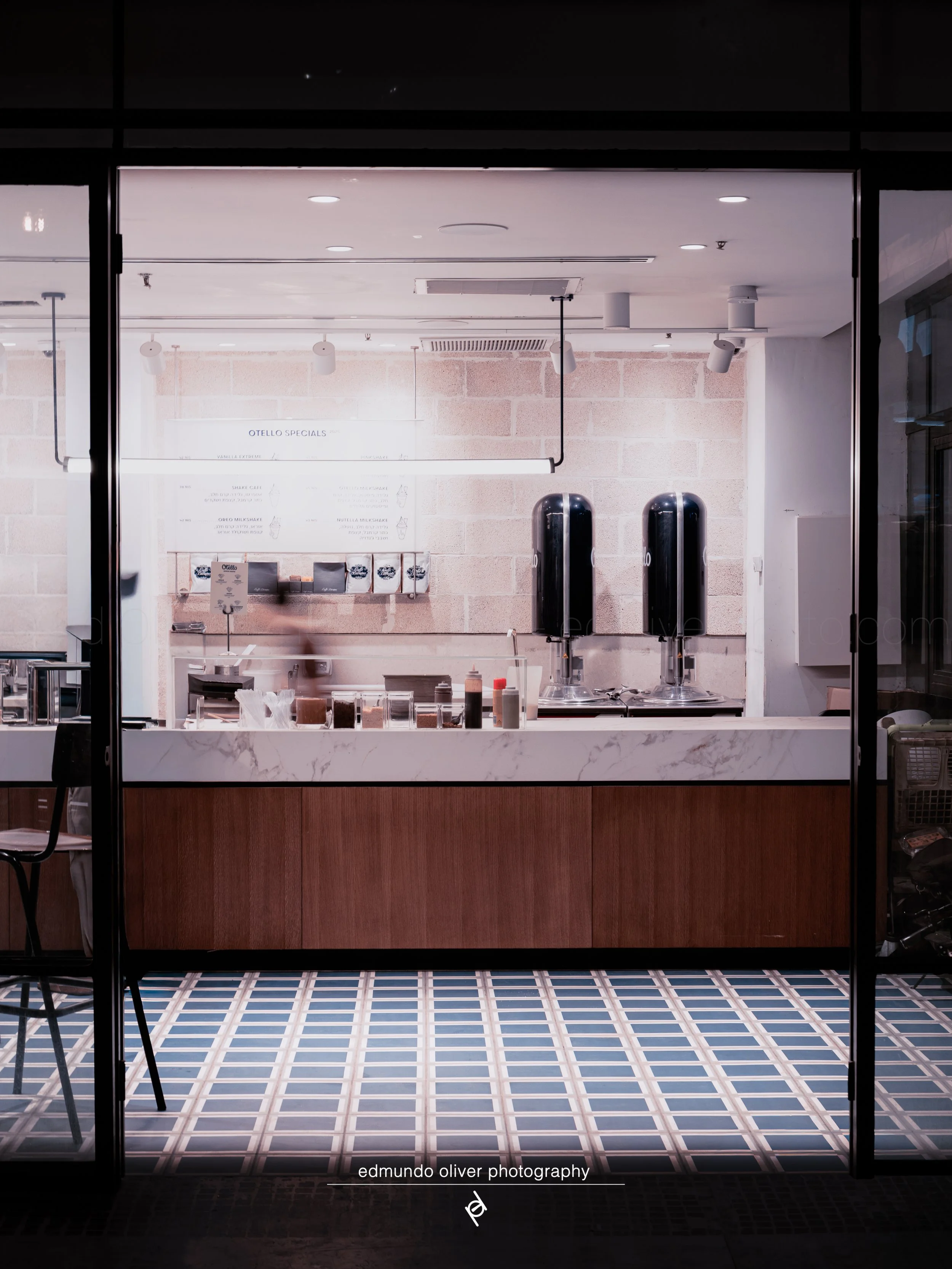 The image shows a view of the interior of a modern cafe or restaurant seen through a large glass window. Inside, there are tiled floors with a geometric pattern, wooden lower cabinets, white walls with a white brick accent wall, and a menu board that