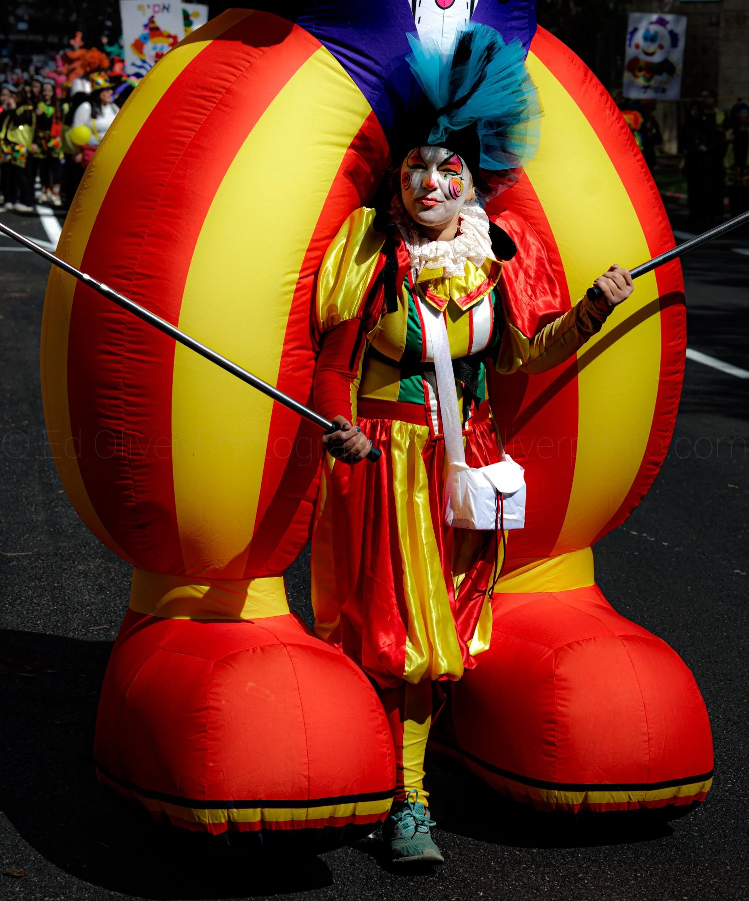 Person dressed as a clown with face paint, wearing colorful costume, standing inside of a large, inflatable, yellow and red armchair, at a parade or festival with onlookers in the background.