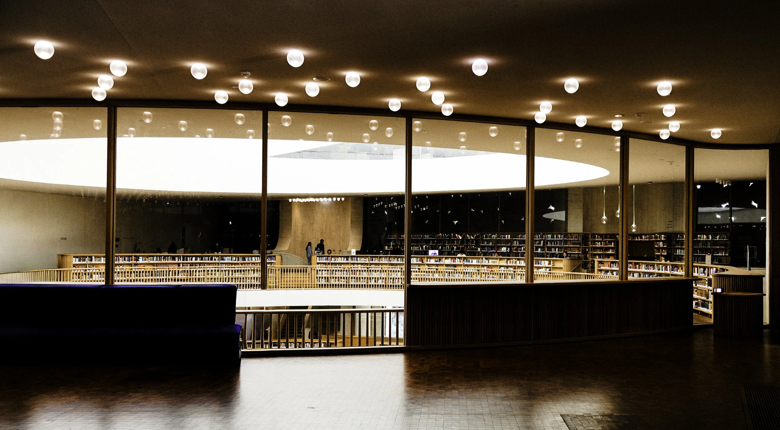 Interior of a modern library with wooden bookshelves, a balcony with glass walls, and circular ceiling lights.