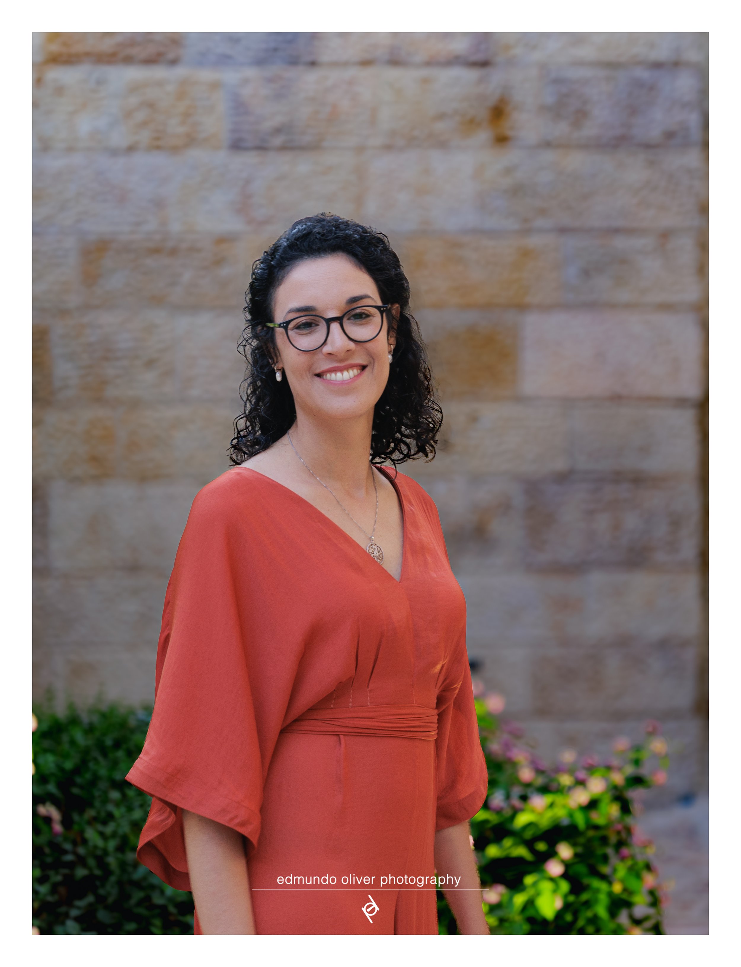 Portrait of a woman with curly dark hair, wearing glasses and a red-orange dress, standing outdoors in front of a stone wall and colourful flowers.