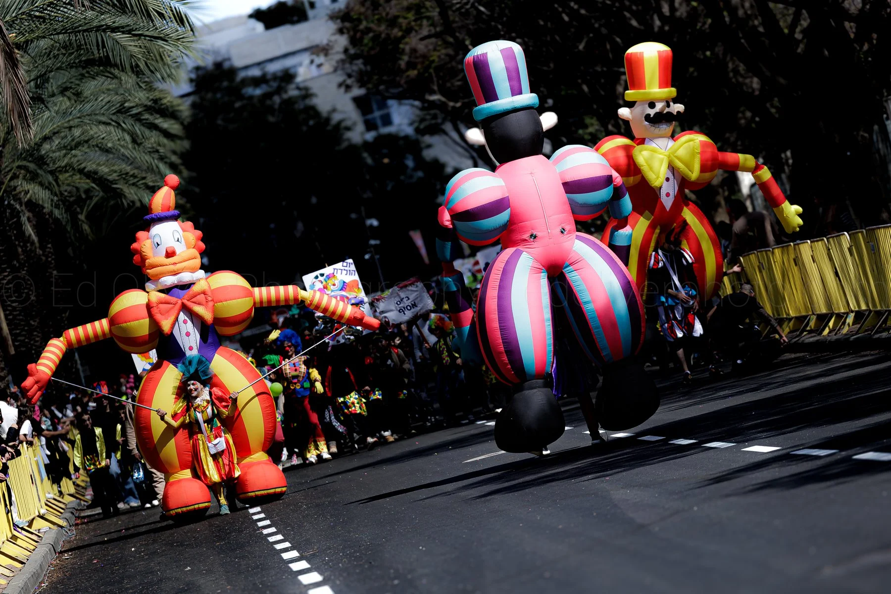 Colorful parade with large clown and jester inflatables, balloons, and performers dressed as clowns on a city street with yellow barriers and spectators.