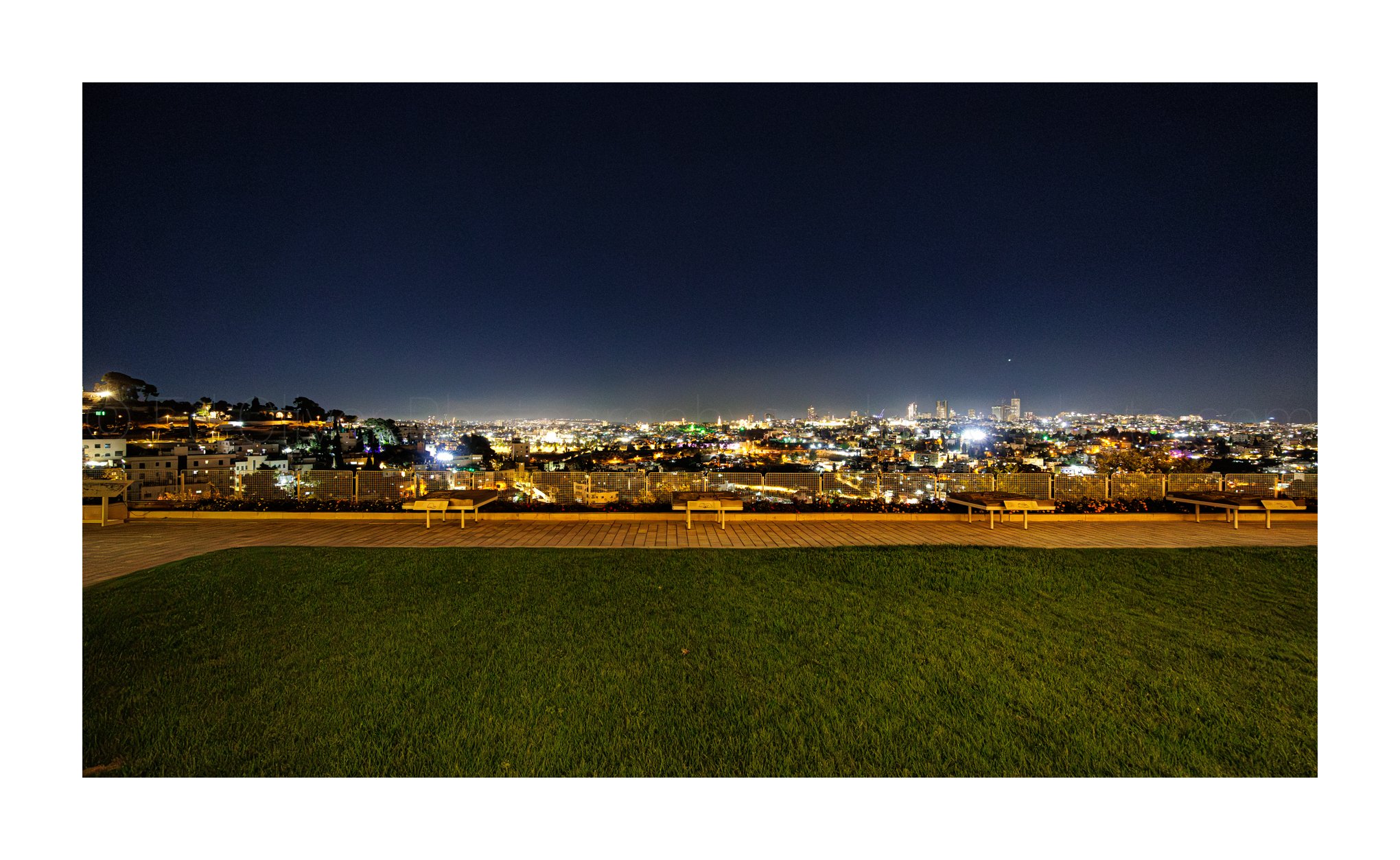 Nighttime cityscape with lit-up buildings, a hill with houses on the left, and a park with benches in the foreground.