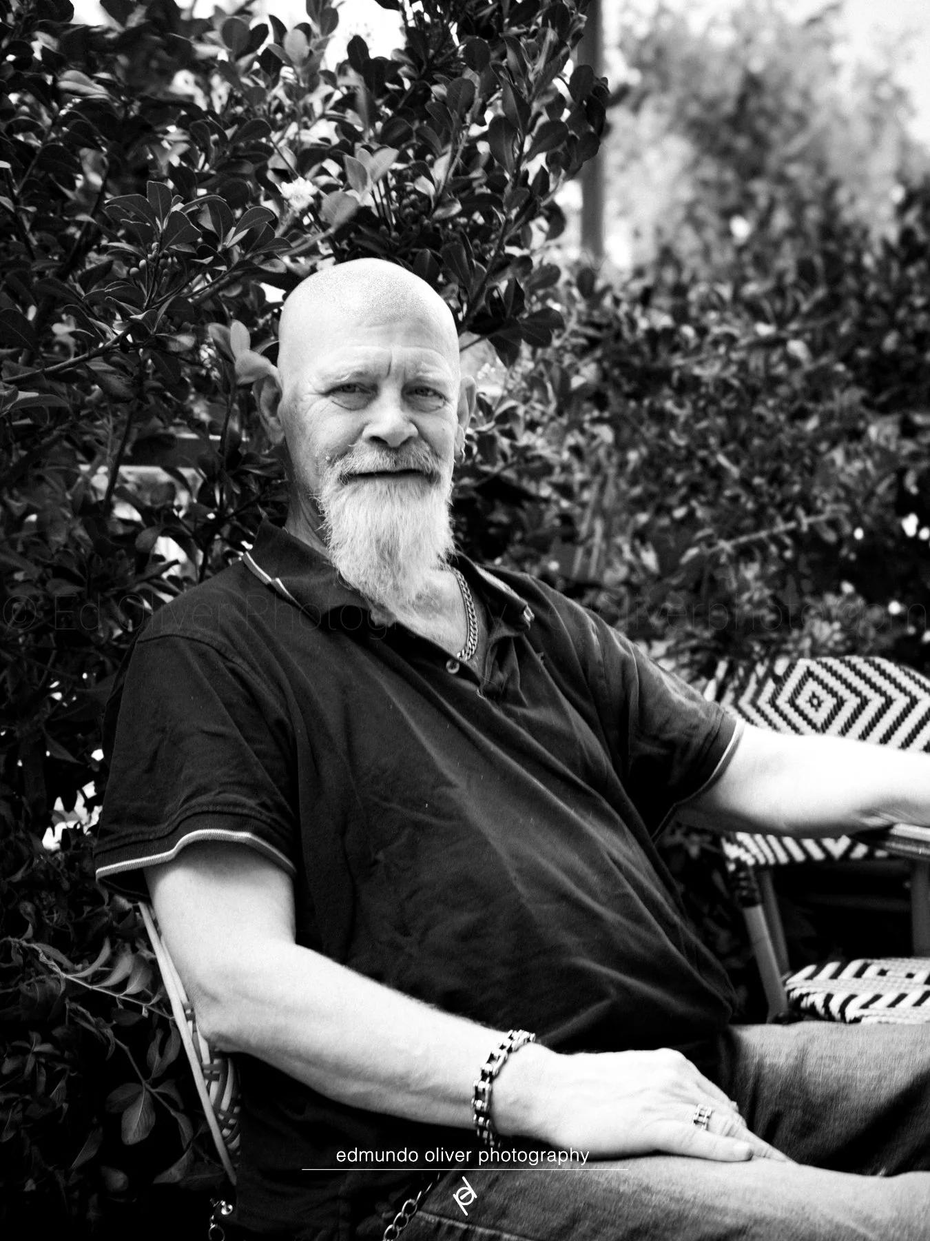 A black and white photo of a man with a beard and shaved head, sitting outdoors among foliage and a patterned chair, smiling slightly at the camera.