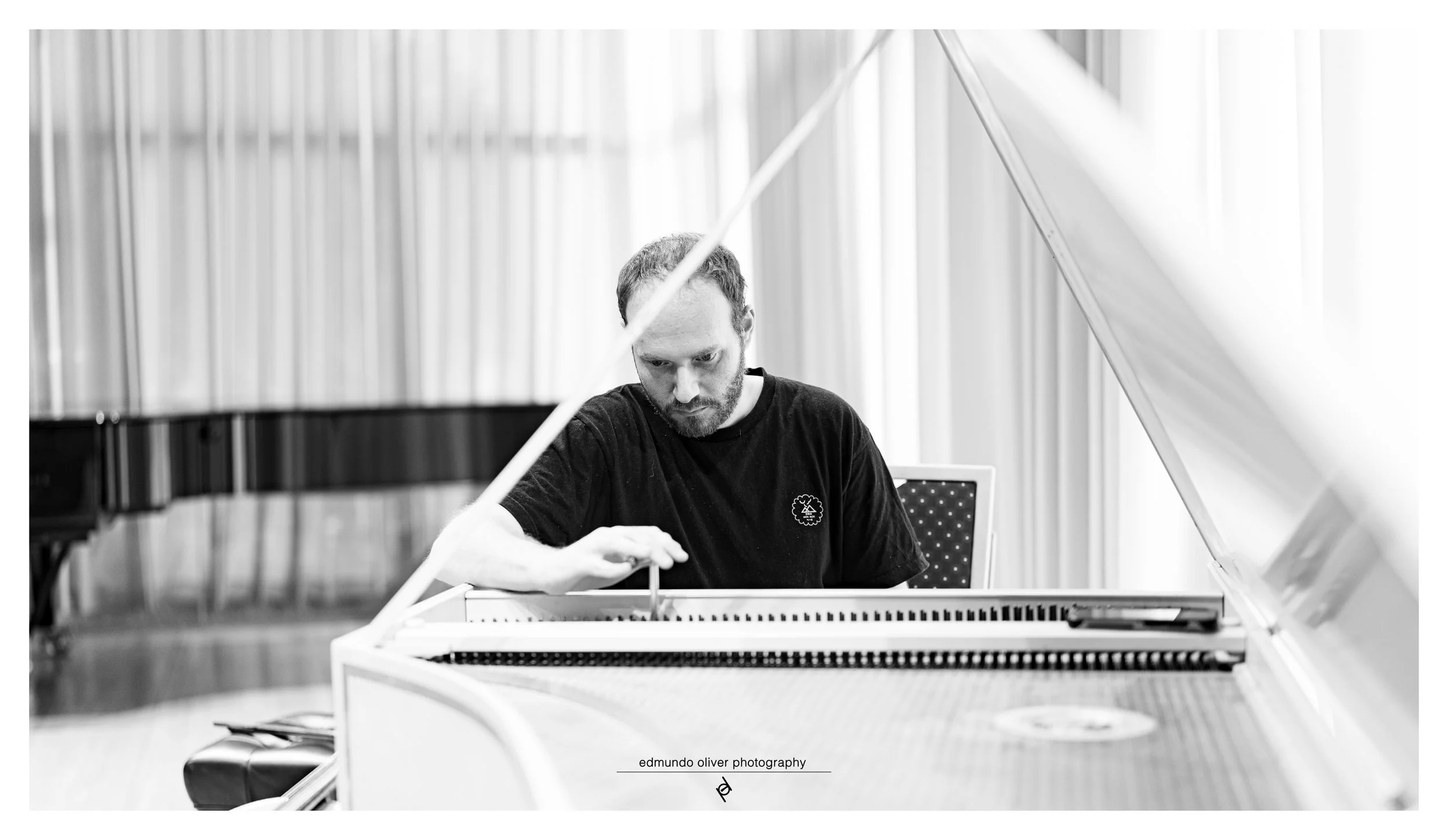 Portrait of a man with a beard and short hair, wearing a black t-shirt,  tuning or adjusting a harpsichord in an indoor space with light-colored curtains or blinds in the background, in Black and White