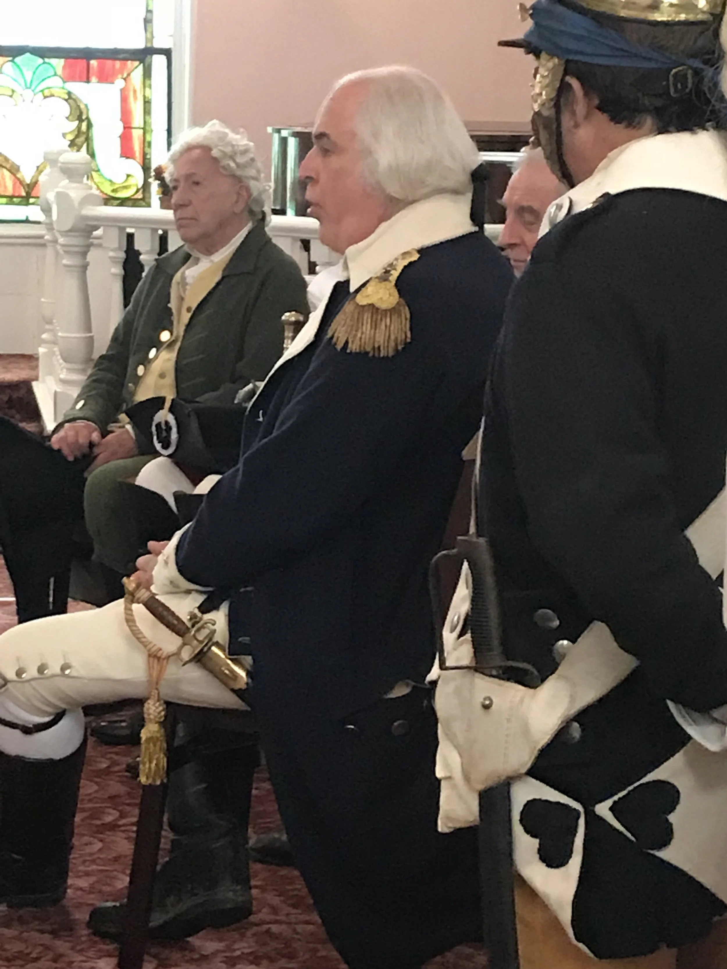 Historical reenactors dressed in colonial military uniforms sitting in a church with stained glass windows.