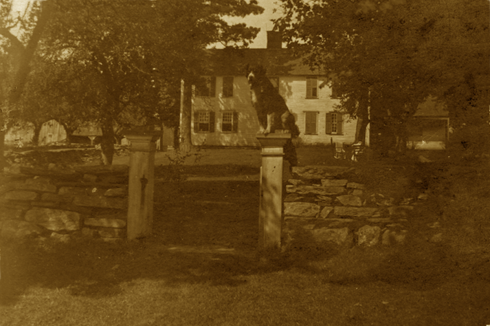 Old sepia-toned photograph of a house with a statue on a pedestal in a yard surrounded by trees and stone walls.
