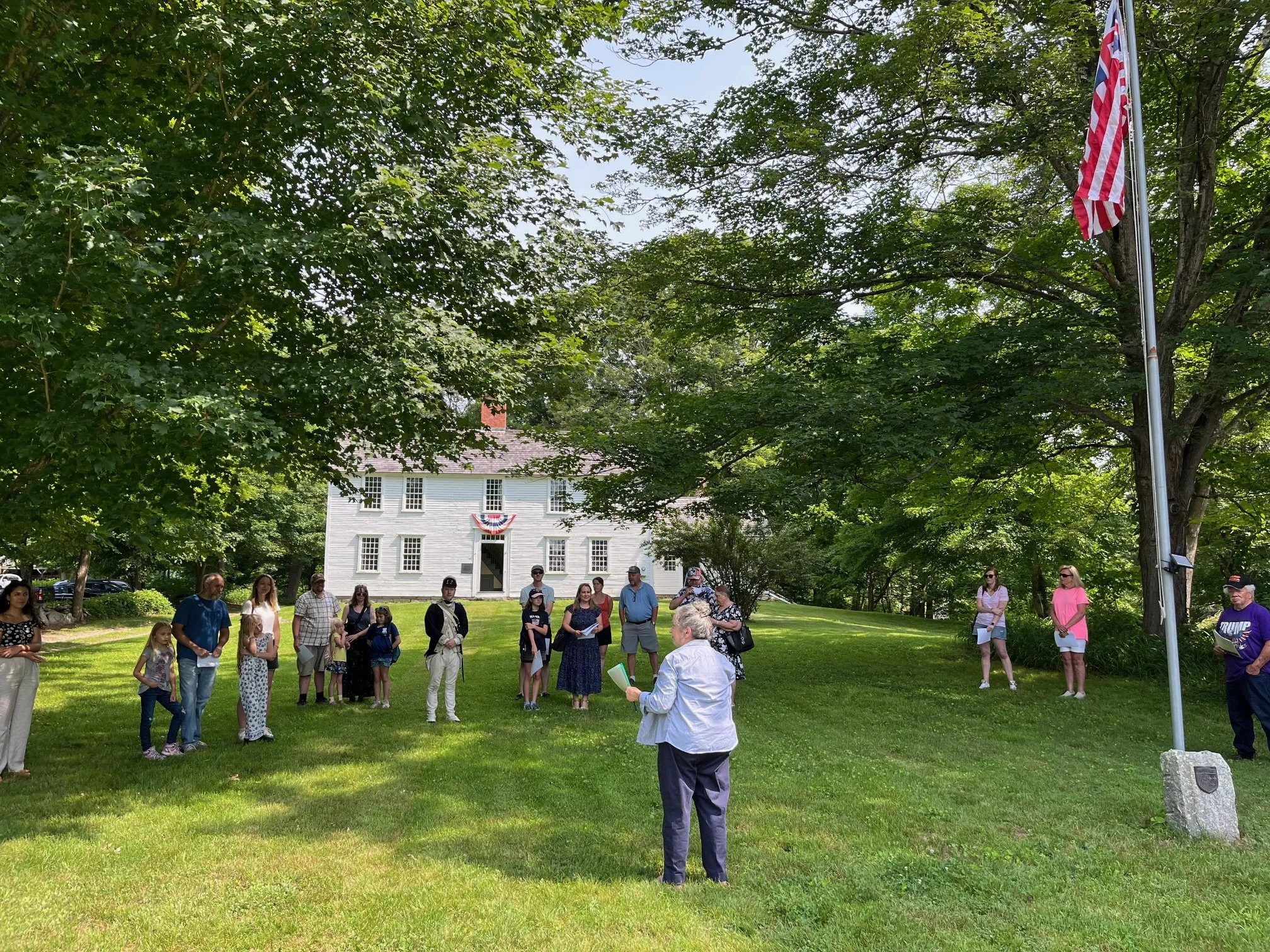 Group of people gathered in a grassy area with trees, a large white house in the background, and an American flag on a flagpole.