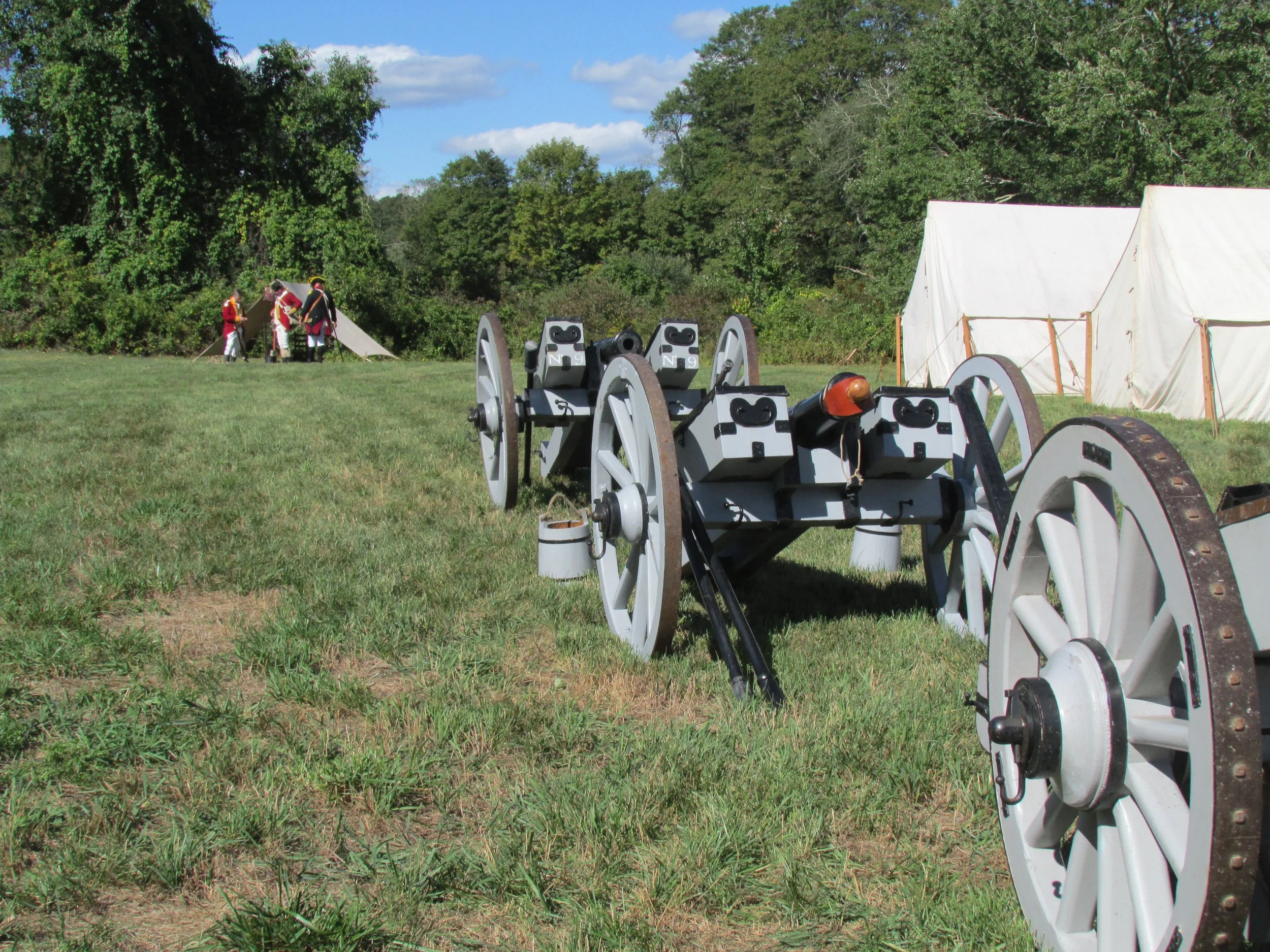 Cannon artillery piece on grass with tents and soldiers dressed as historical figures in the background.