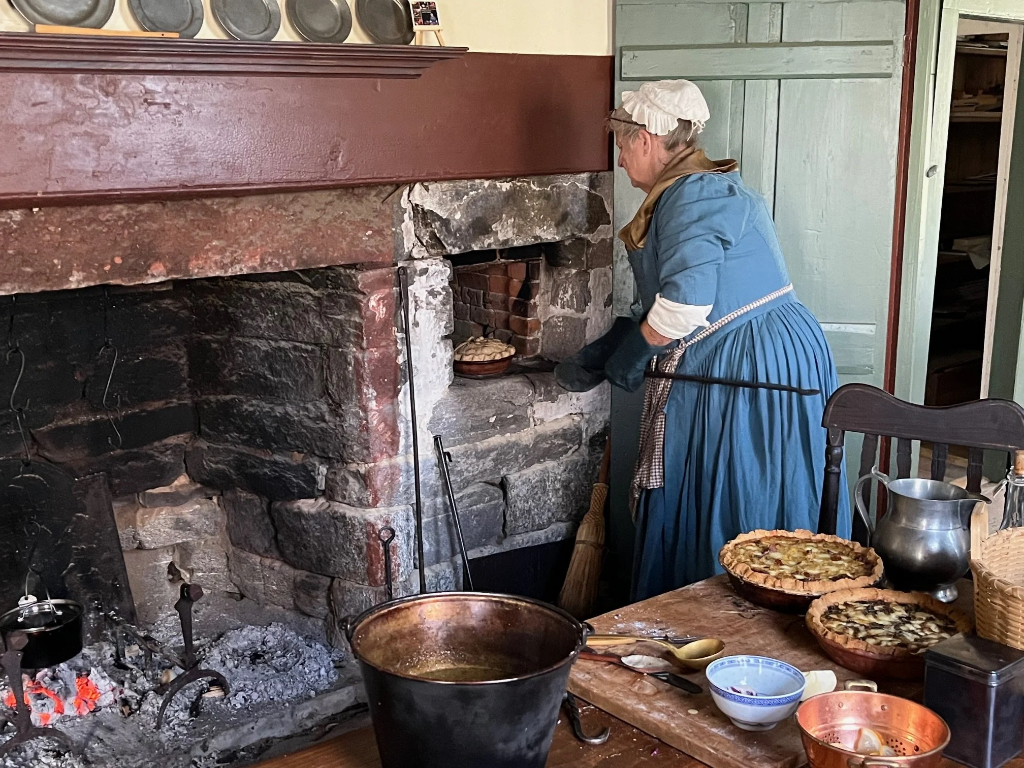 A woman dressed in historical clothing, including a bonnet and long dress, is baking bread in an old-fashioned brick oven. There are two pies on the table in front of her, along with various baking utensils and a metal pitcher.