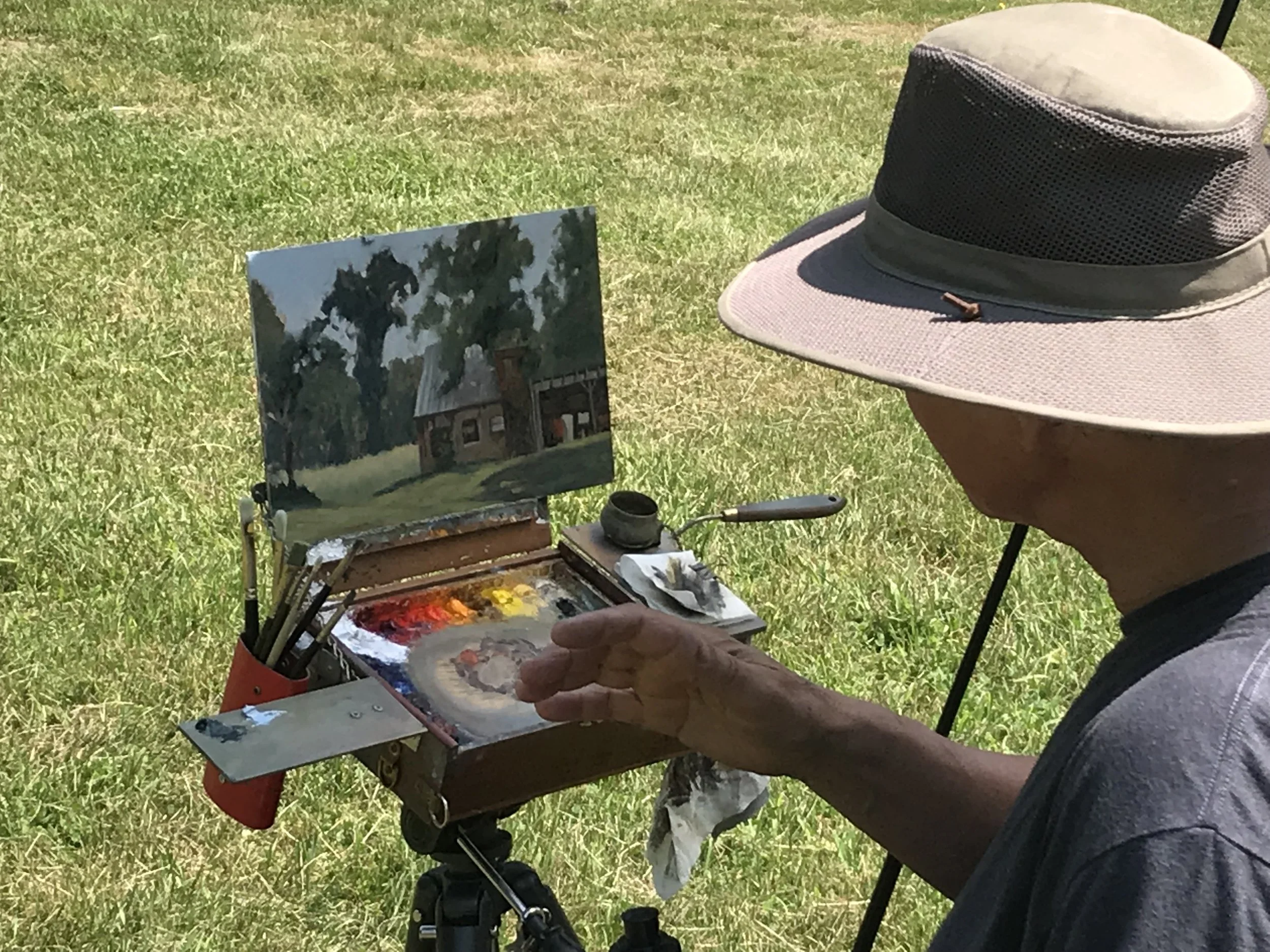 An artist painting a landscape scene on a small portable easel outdoors, wearing a wide-brimmed hat, with a palette of paints and brushes, and a finished painting of a house surrounded by trees in the background.