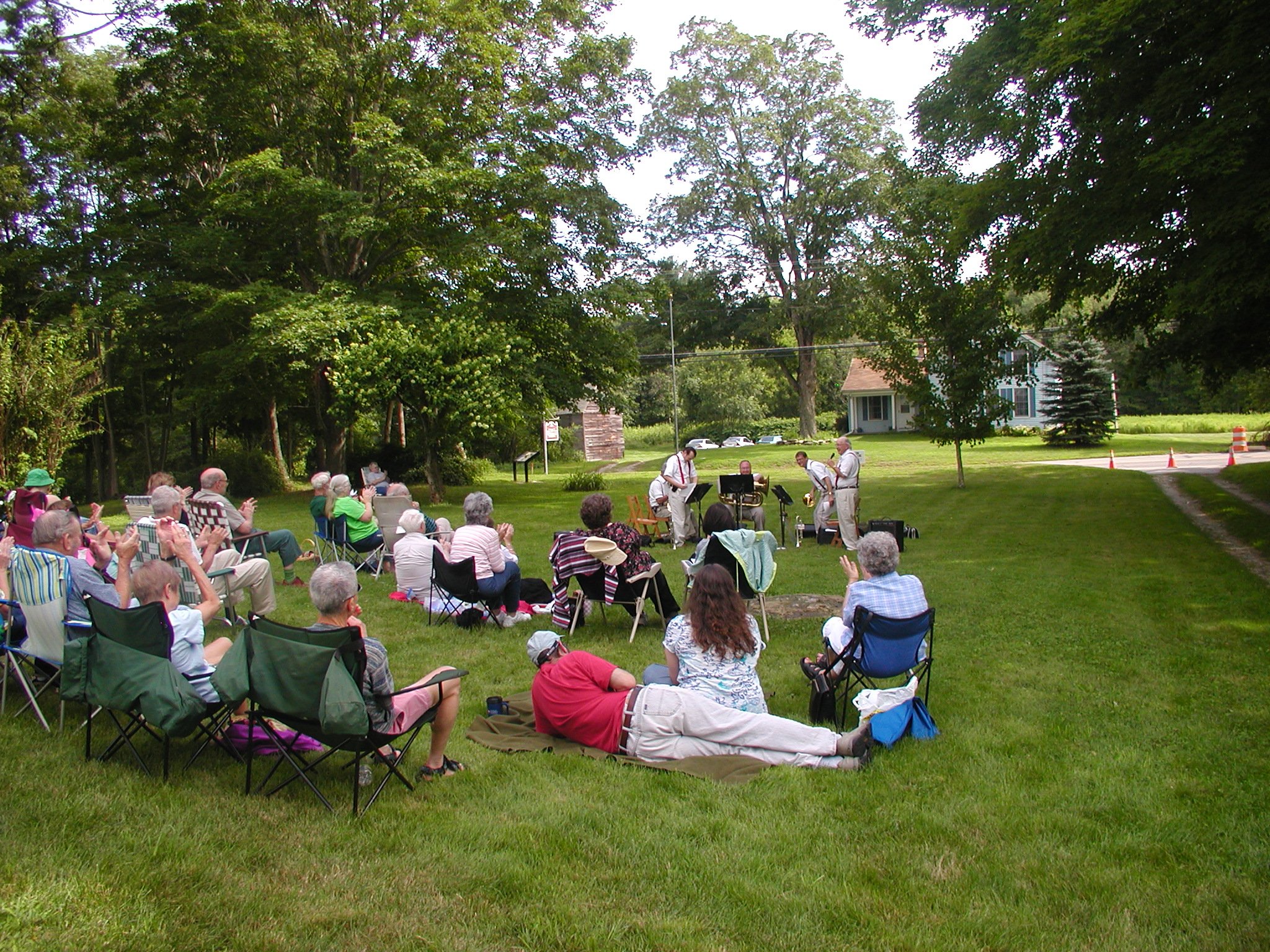 An outdoor concert in a park with a small audience seated on lawn chairs and blankets, watching a band perform on a grassy area under trees.