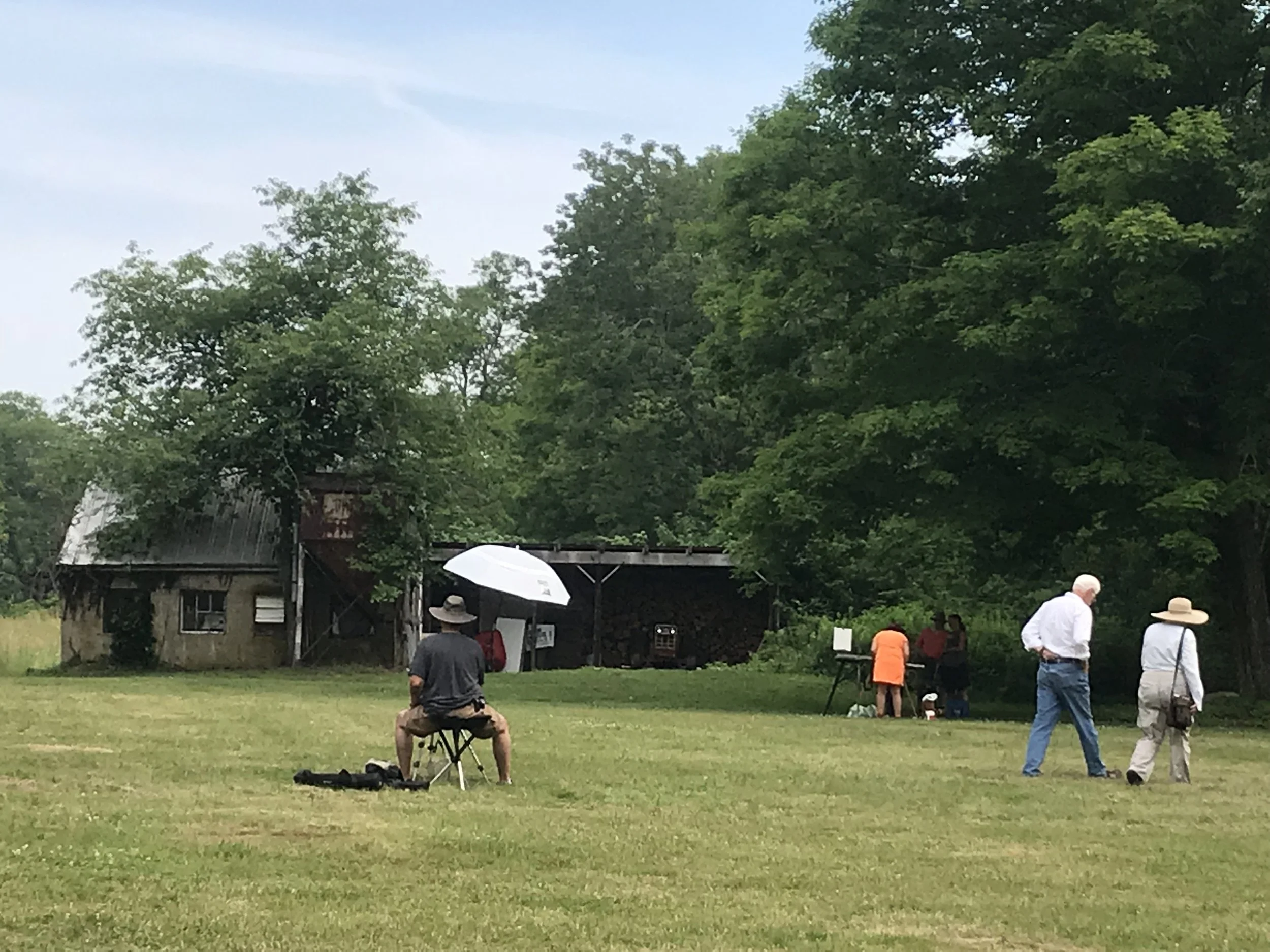 People gathered outdoors on a grassy field with trees and an old barn in the background. Some individuals are standing, while others are seated or sitting under umbrellas, appearing to be engaged in a small event or gathering.