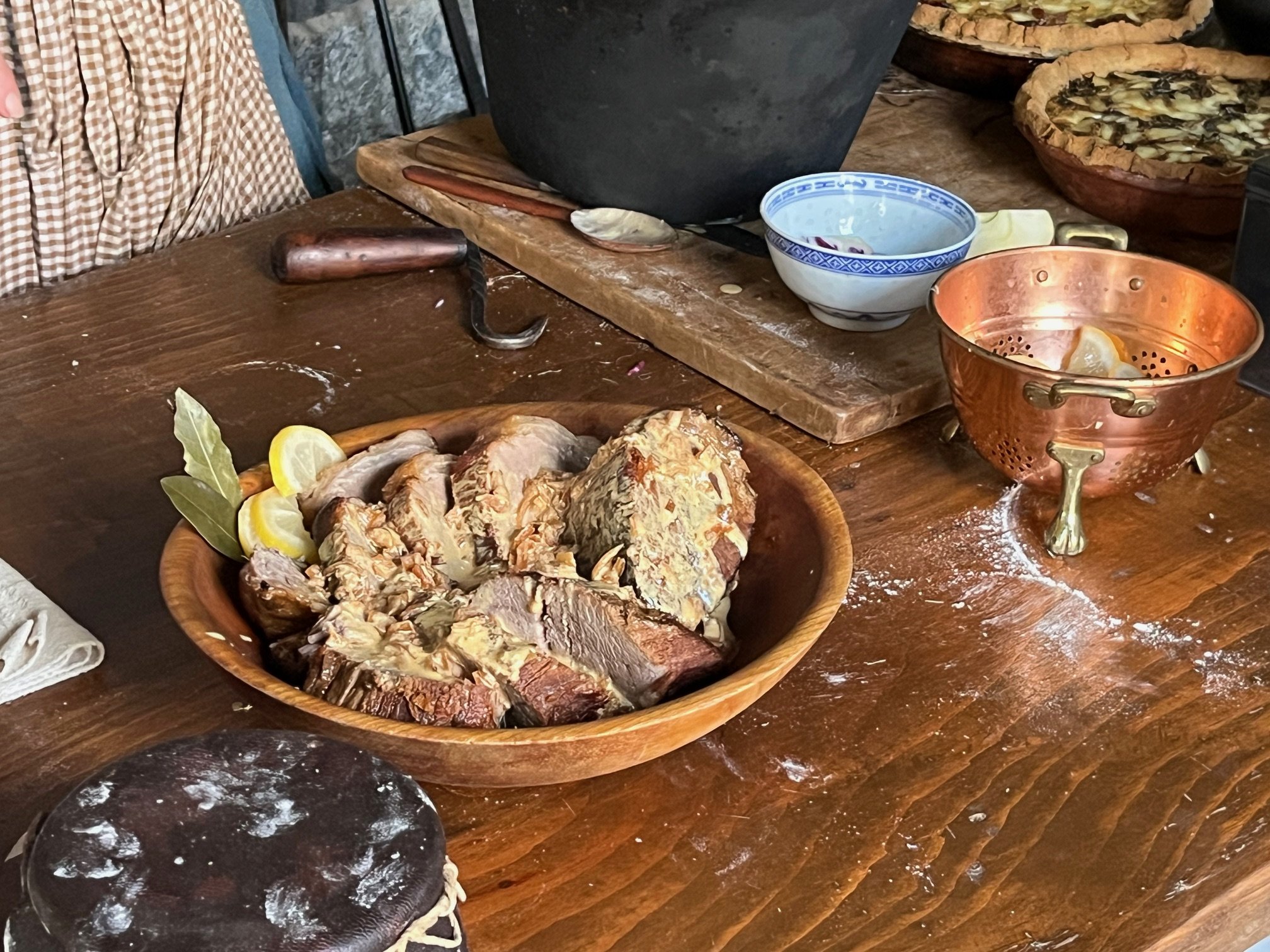 A wooden table with a large wooden bowl of cooked meat garnished with lemon wedges and herbs, a copper bowl with lemon slices, a white and blue bowl, and a black pot. Some kitchen utensils and ingredients are also visible.