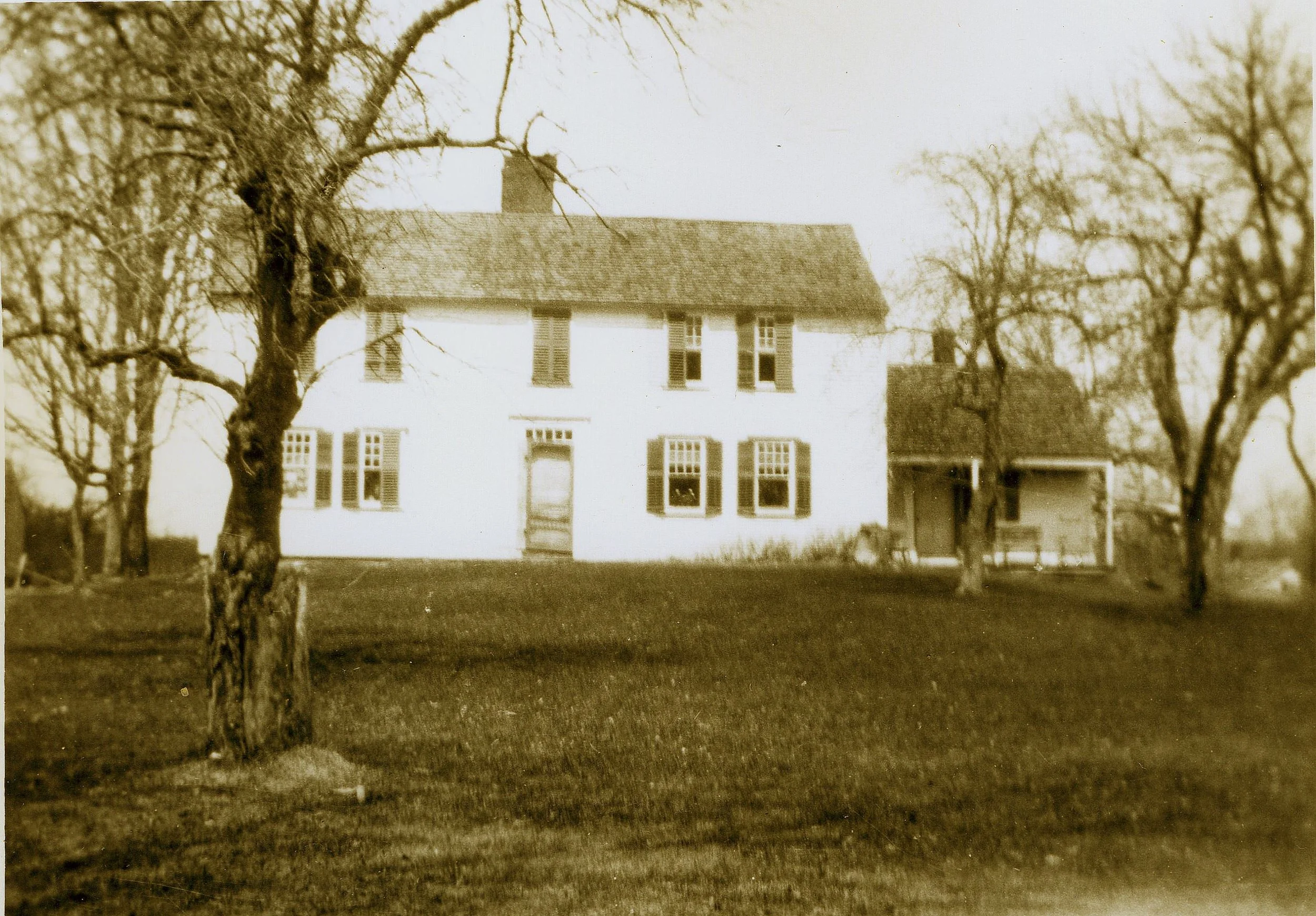 An old sepia-tone photograph of a two-story white house with multiple windows, a central door, and trees in the front yard.