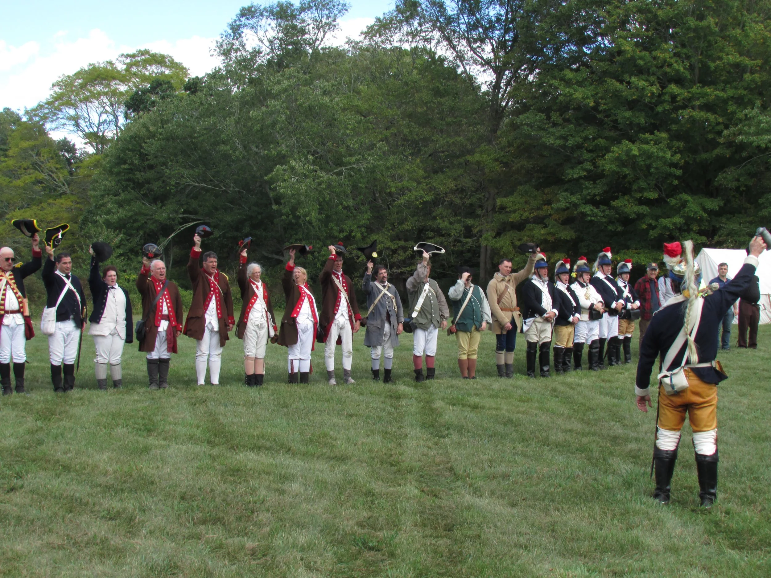Group of people dressed in historic military uniforms, participating in a reenactment on a grassy field with trees in the background.