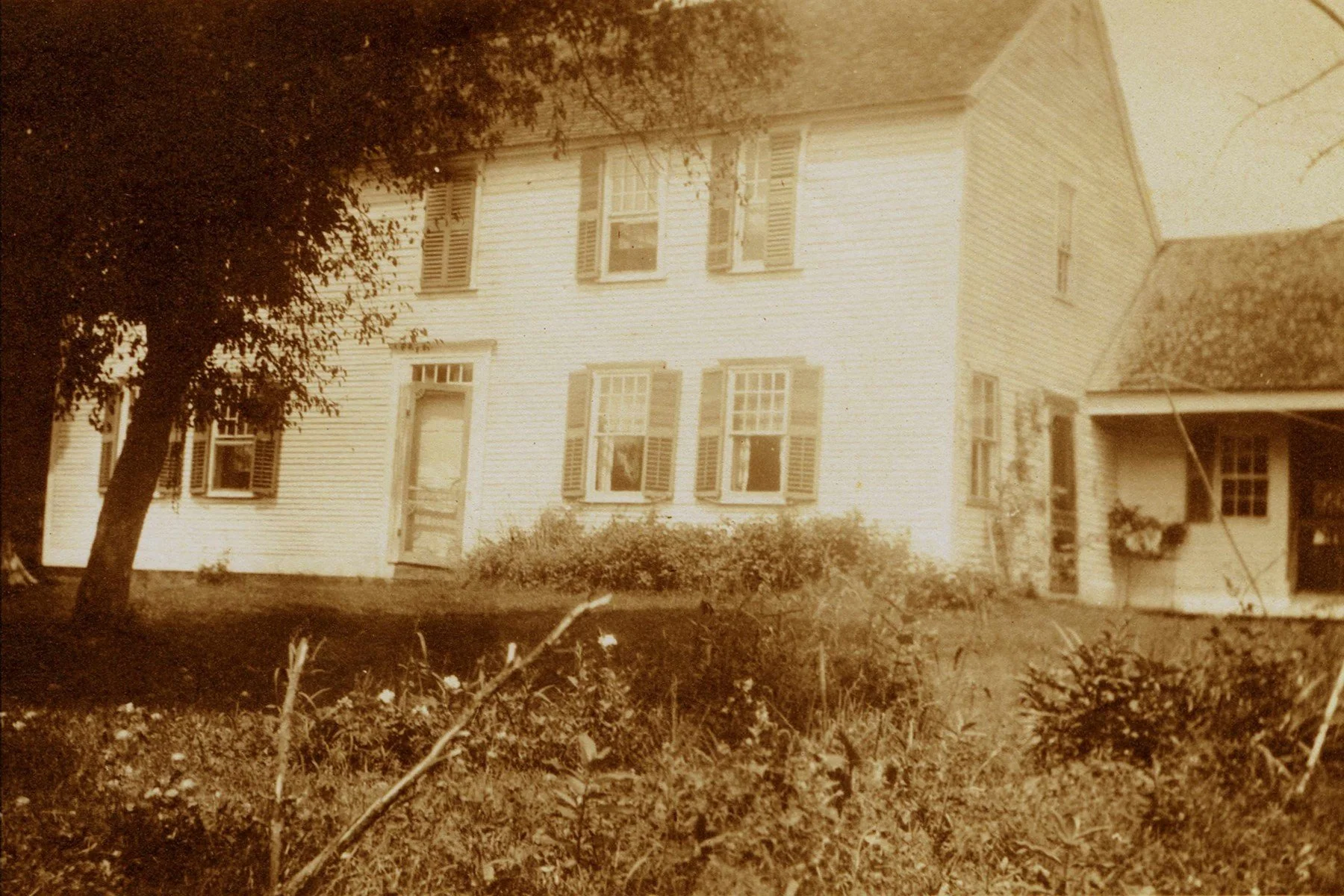 An old sepia-toned photograph of a two-story house with multiple windows and shutters, surrounded by a garden with bushes and a tree to the left.