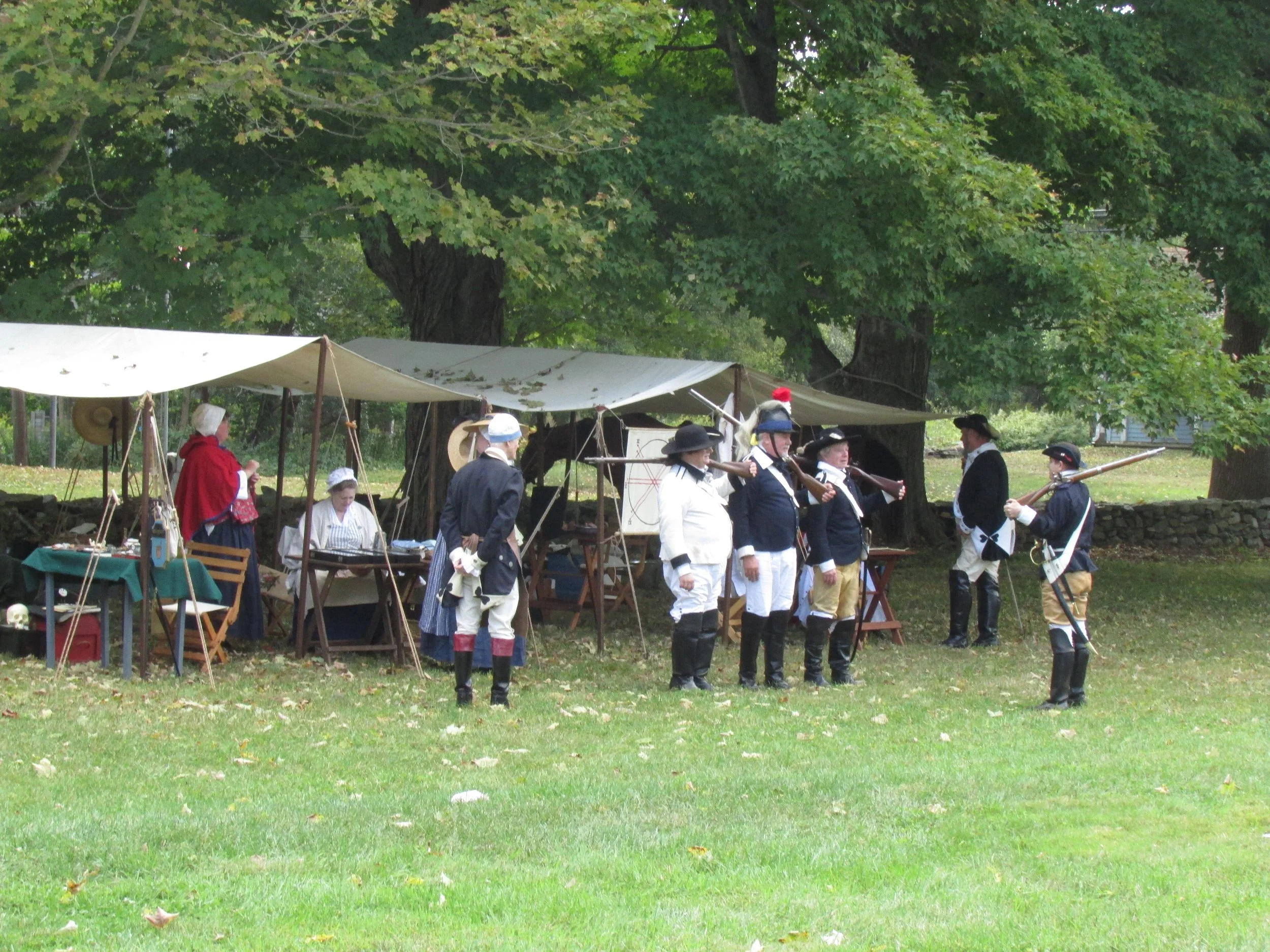 Reenactment of a historical military scene with people dressed in 18th-century military uniforms, some holding muskets, set in a park with trees and tents.