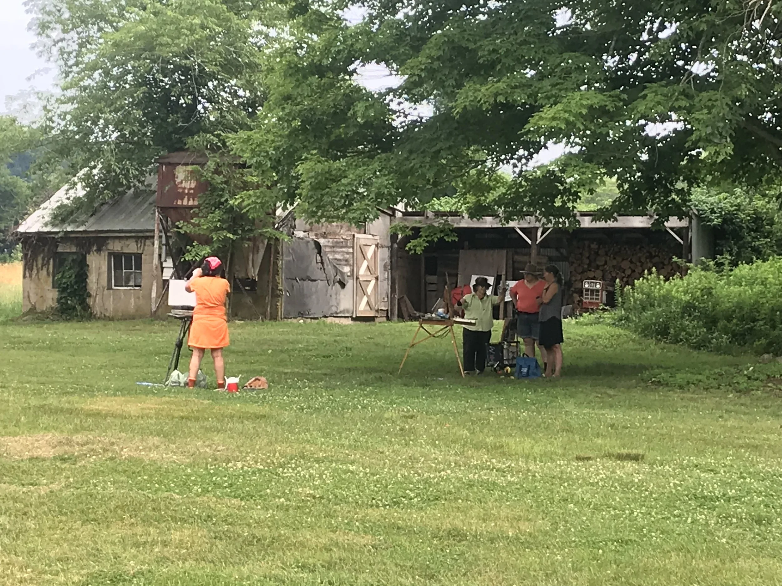 A group of people is gathered outdoors near an old barn, with green grass and trees surrounding them. One person is operating camera equipment, and others are conversing or standing near tables and equipment.