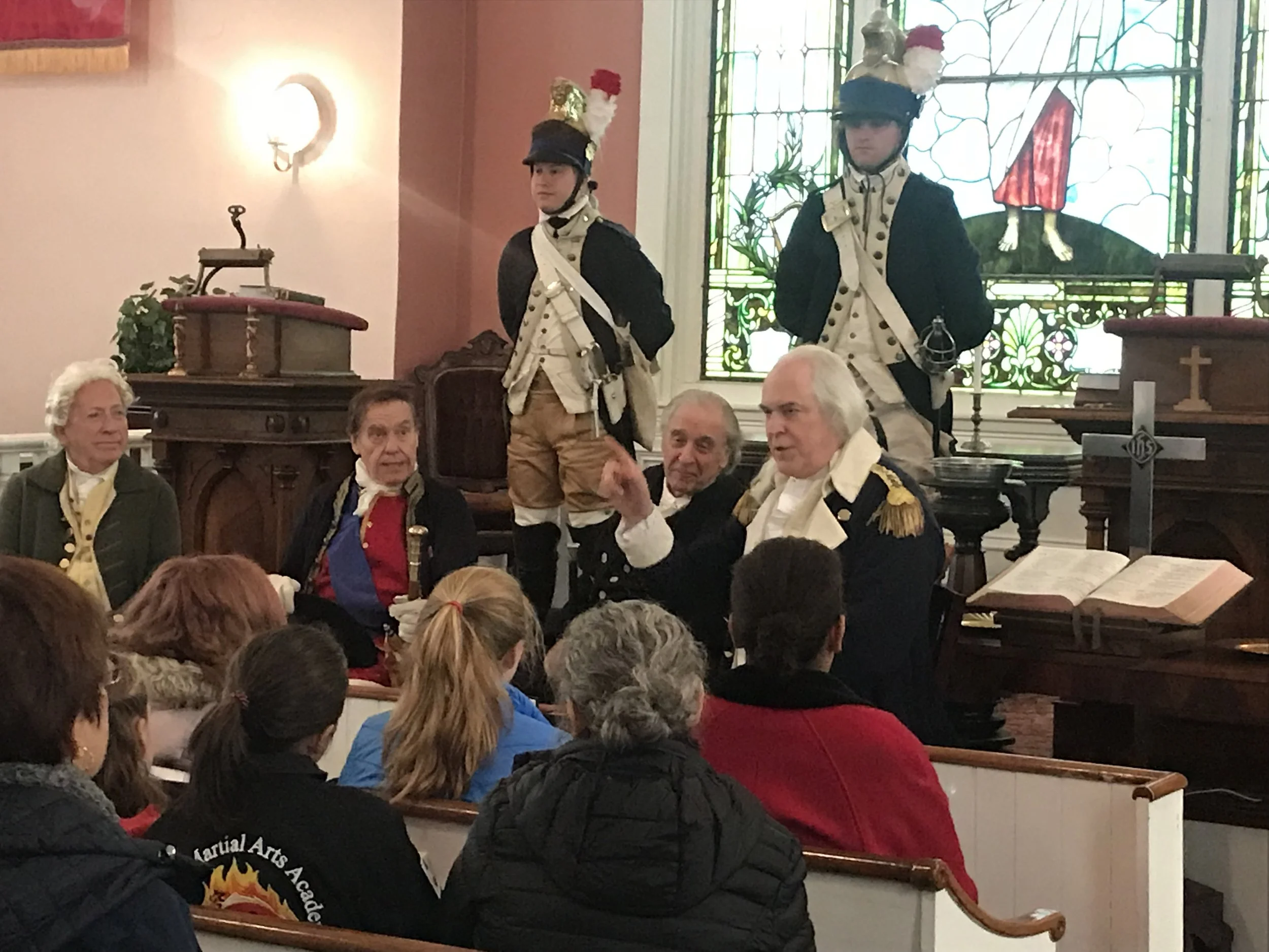 A historical reenactment inside a church with men in Revolutionary War uniforms speaking to an audience. The scene includes a stained glass window, a wooden pulpit, and a cross on an open book on a stand.